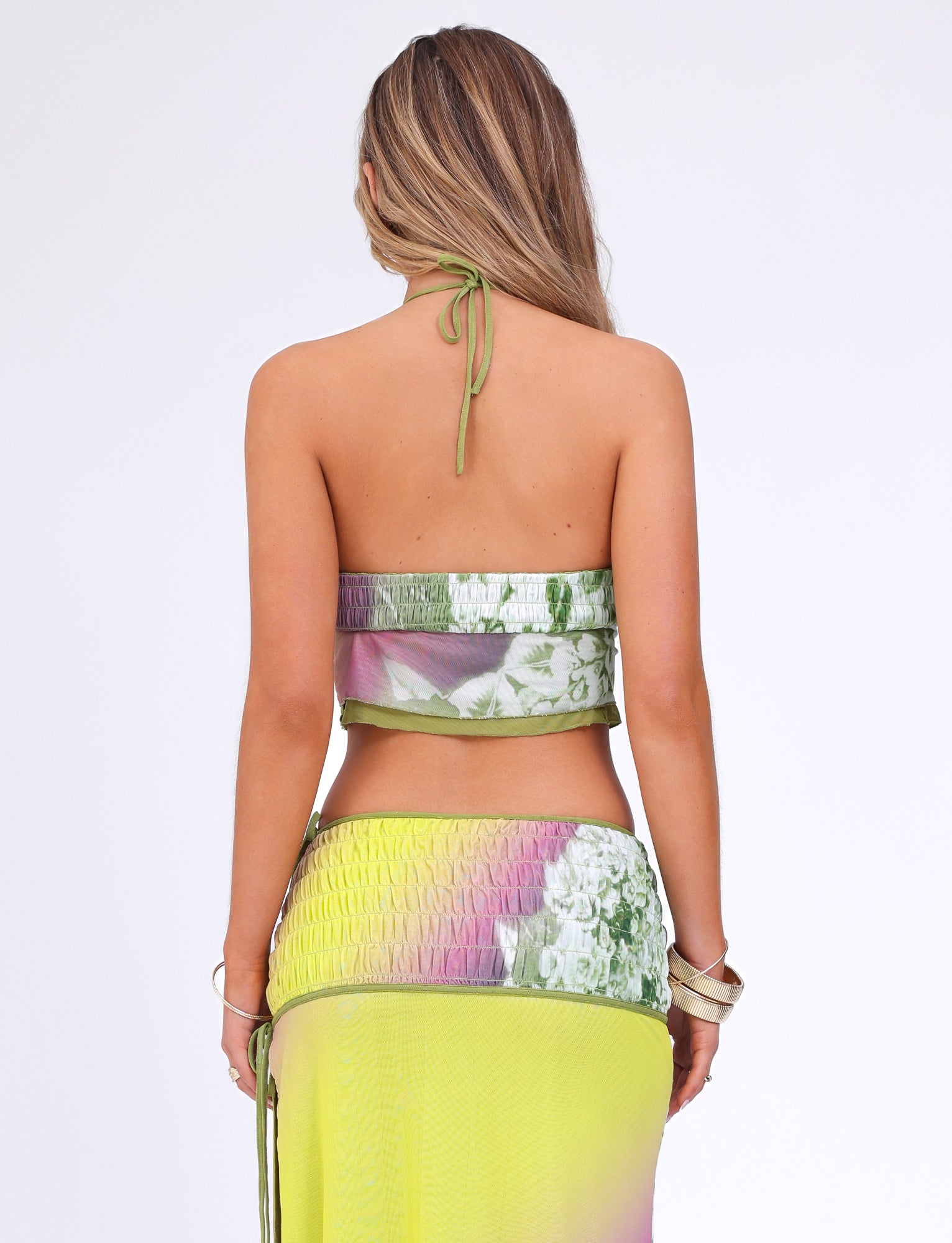 A person with long hair stands with their back to the camera. They are wearing the TIGERMIST WAVERLY TOP - GREEN : FLORAL : STRIPE, a crop-length halter top paired with a matching skirt in a green, pink, and yellow floral pattern. The stretchy fabric outfit exposes their back. The background is plain white.