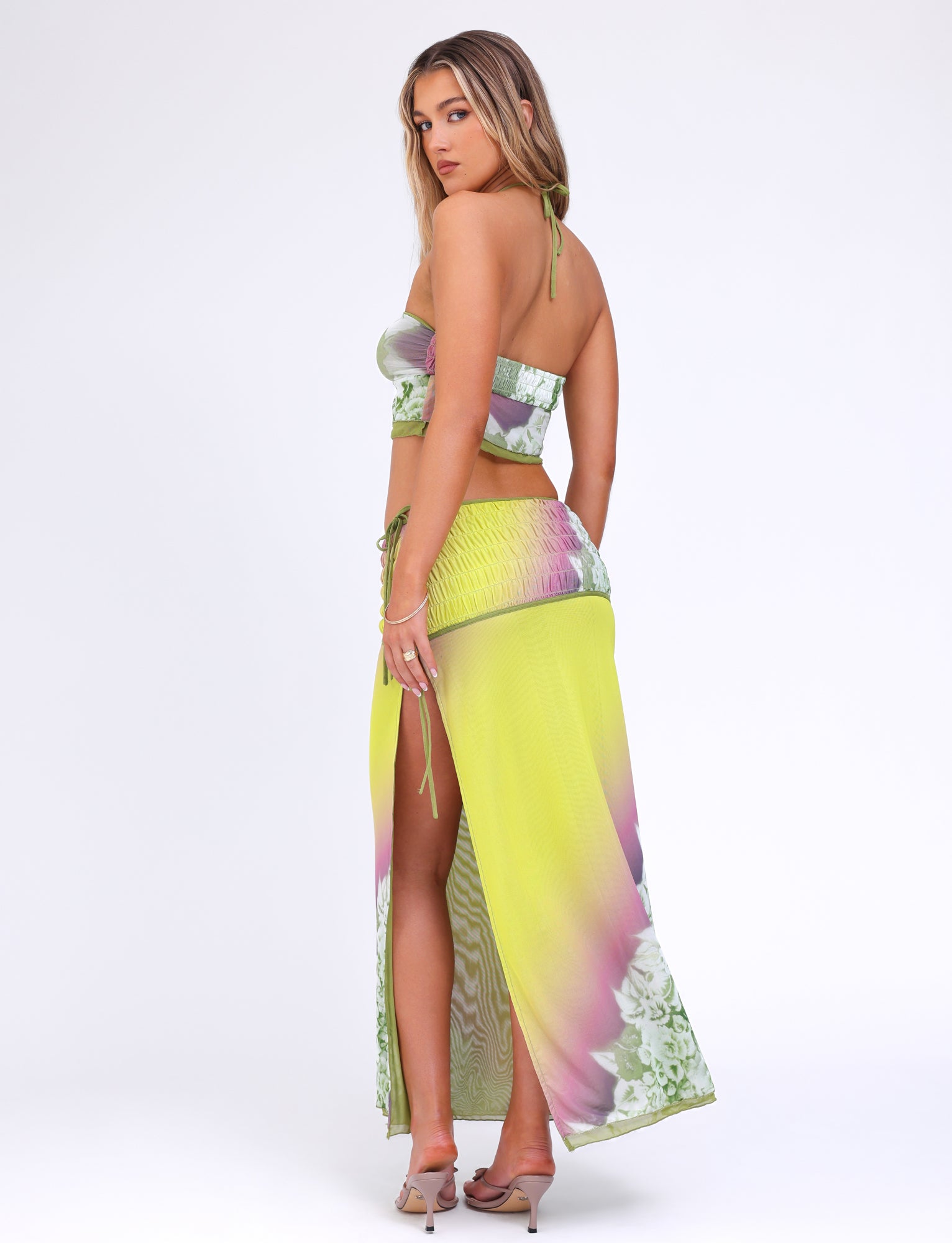 A woman standing against a plain white background is wearing a two-piece outfit from TIGERMIST, featuring the WAVERLY SKIRT in green with floral and stripe patterns. The ensemble includes a green and purple tie-dye crop top paired with the matching high-waisted, ankle-length reversible skirt with side slits. She has long, wavy hair and is wearing high-heeled shoes.