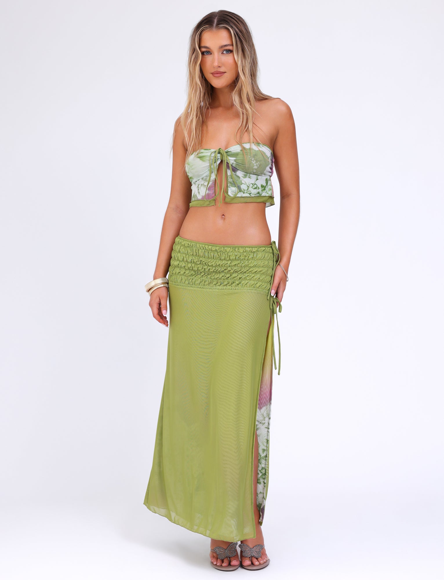 A woman with long, blonde hair is wearing the TIGERMIST WAVERLY SKIRT - GREEN : FLORAL : STRIPE and a matching green floral bandeau top with a slit. The mesh fabrication of her outfit adds an elegant touch as she stands on a white background, accessorized with bracelets on her wrists.