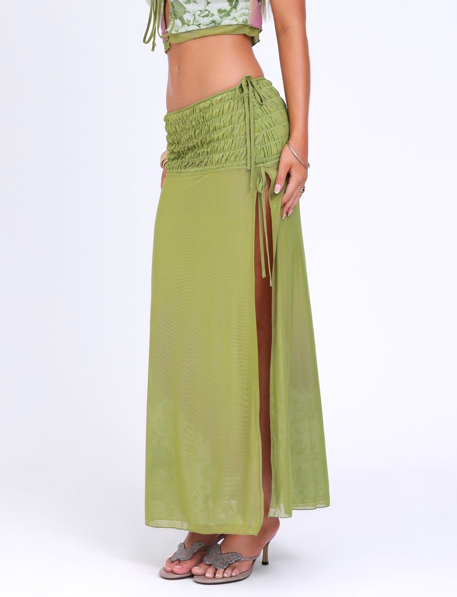 A person wearing the WAVERLY SKIRT - GREEN : FLORAL : STRIPE by TIGERMIST, a sheer green maxi skirt with a side slit and gathered detailing at the low-rise waist, paired with a matching cropped top and flip-flop sandals, stands against a white background. The person poses with one arm resting on the side, displaying their hand adorned with rings.