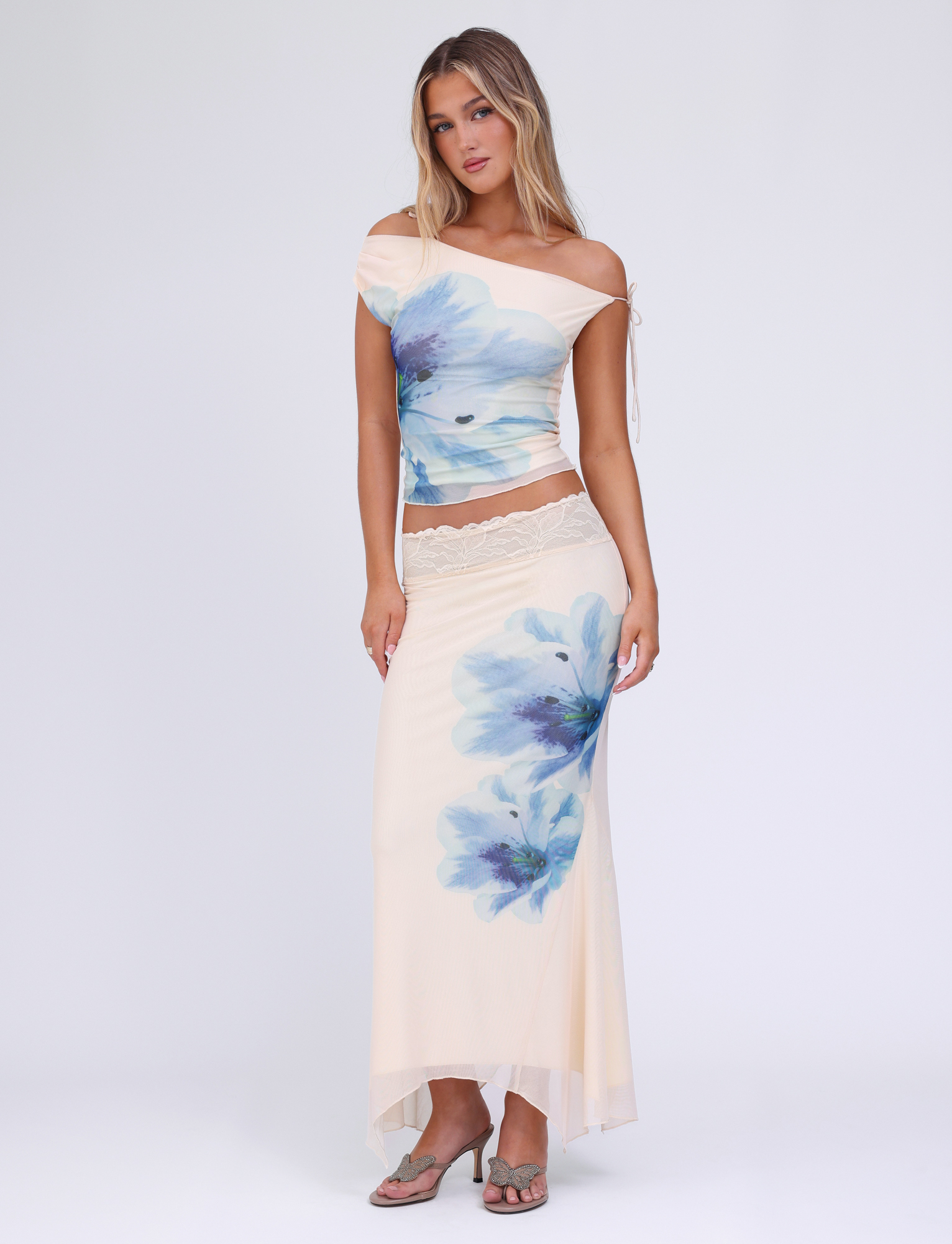 A woman with long, loose hair poses against a plain white background wearing the TIGERMIST LILLIAN TOP in cream floral with an off-shoulder mesh design, paired with a matching maxi skirt featuring blue prints and a lace waistband.