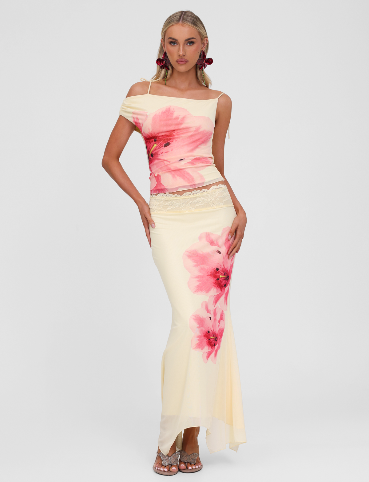 A woman poses in the TIGERMIST LILLIAN TOP - YELLOW, featuring mesh fabrication and lace trim at the waist. She pairs it with silver heels and flower earrings, standing against a neutral backdrop.