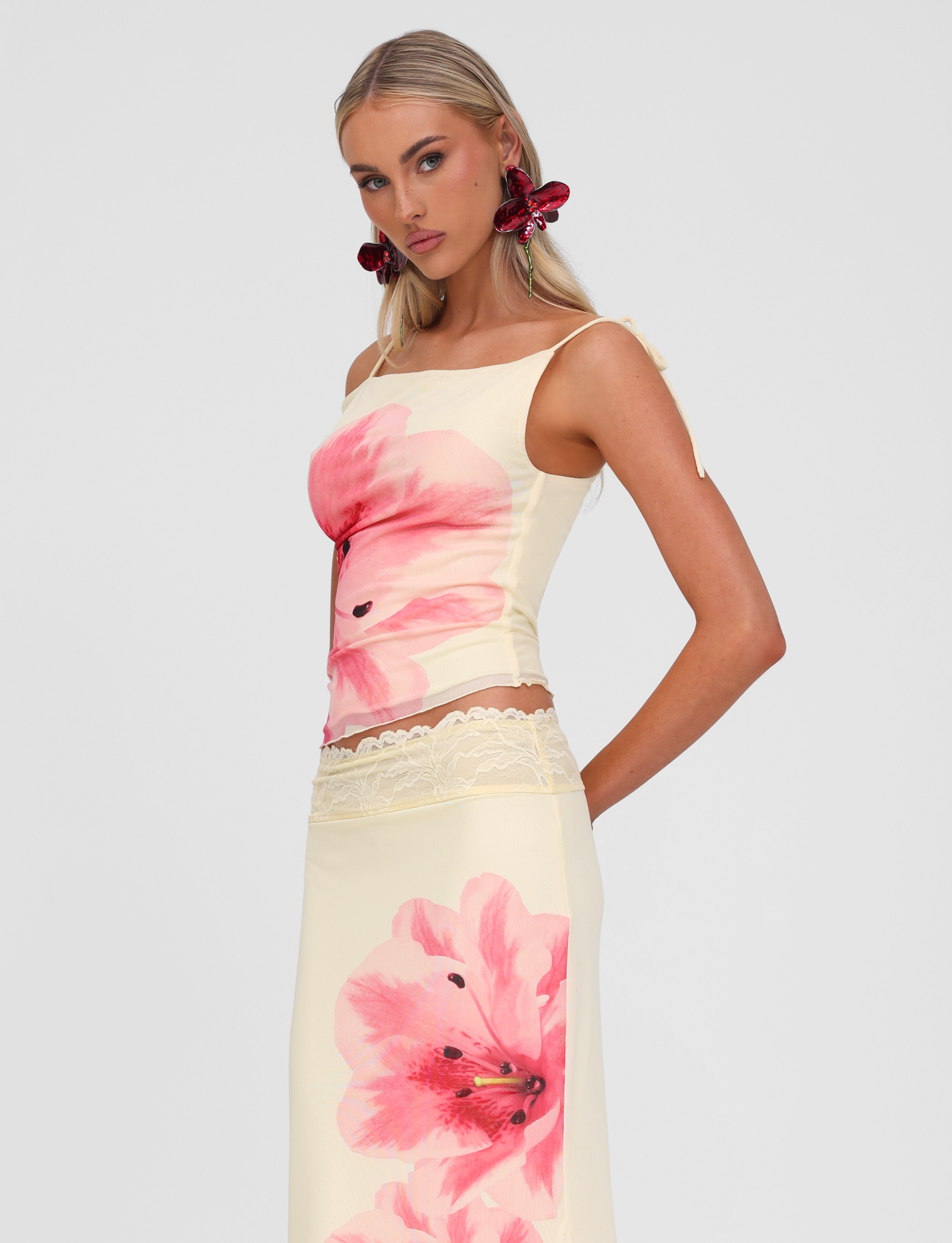 A woman with long blonde hair wears the TIGERMIST LILLIAN TOP in yellow, a fitted off-shoulder style with large pink floral design and lace trim. She pairs it with red flower earrings and poses against a plain light gray background.