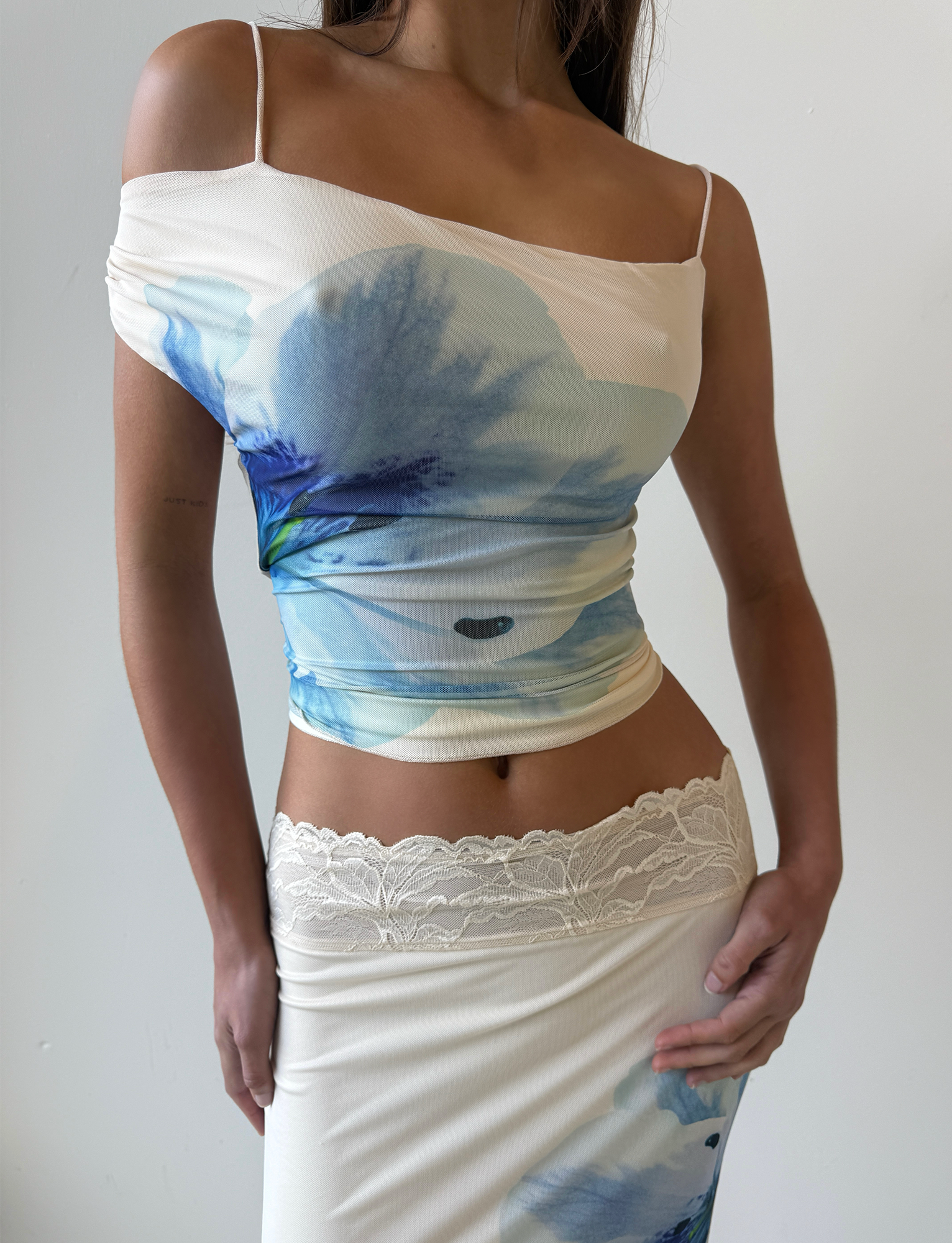 A woman wears the TIGERMIST LILLIAN TOP in cream floral, an asymmetrical off-shoulder style, paired with a matching skirt with lace waistband trim. The background is plain and light-colored.