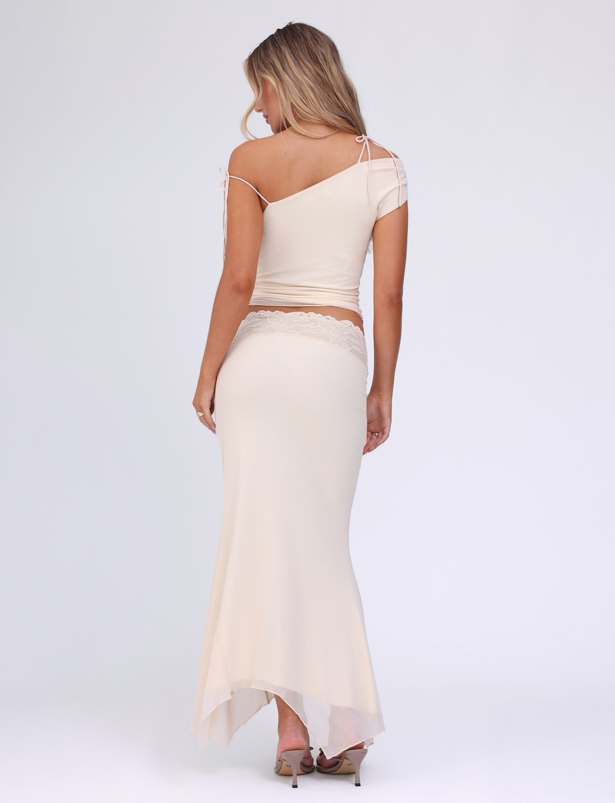 A woman with long blonde hair poses with her back to the camera against a plain white background, wearing the TIGERMIST LILLIAN SKIRT in white, cream, and floral, paired with an asymmetrical top and high heels.