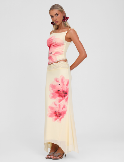 A woman wearing a cream-colored sleeveless top and the TIGERMIST LILLIAN SKIRT in yellow stands against a plain white background, paired with embellished sandals.