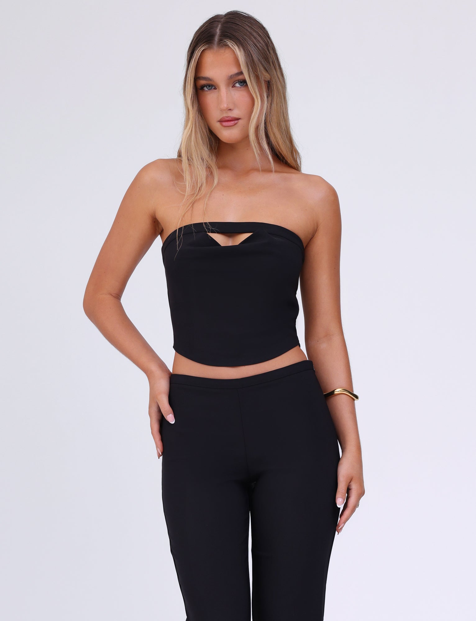A woman with long blonde hair is wearing the TIGERMIST AUDRIE TOP in black, featuring a small cut-out detail at the bust, paired with black pants. She stands with one hand on her hip and the other resting by her side, accessorized with a gold bracelet on her left wrist.