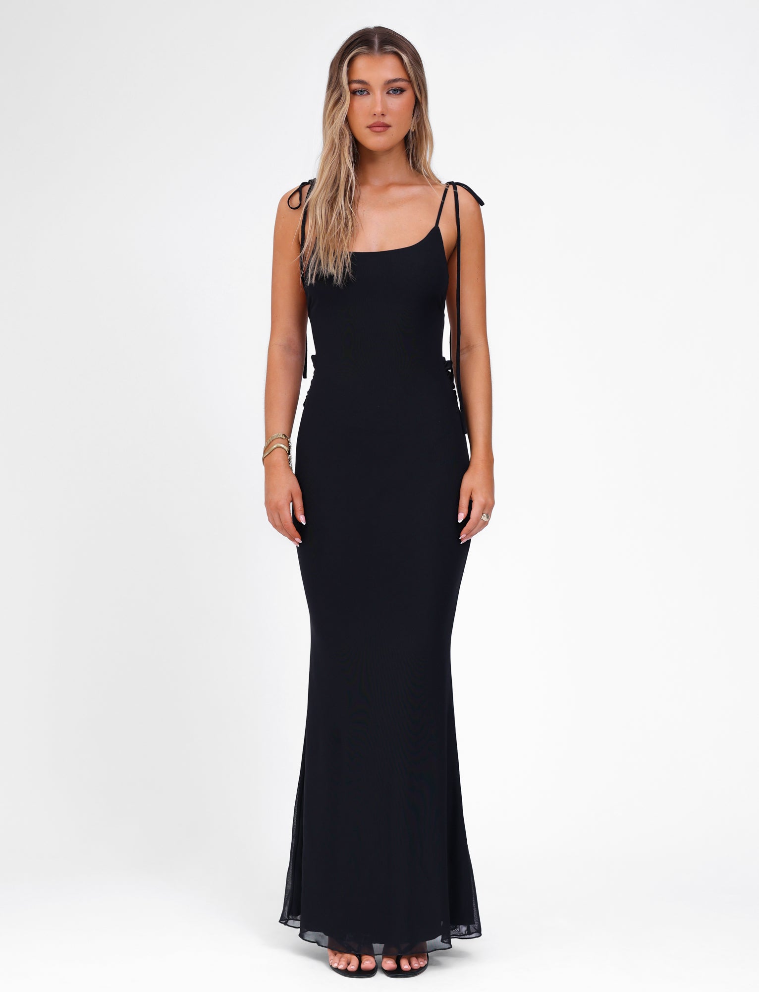 A woman with long blonde hair is wearing the CYRIL DRESS - BLACK by TIGERMIST. This sleeveless, form-fitting dress features adjustable straps and pairs seamlessly with her black high-heeled sandals. She stands against a plain white background, facing the camera with a neutral expression and minimal jewelry on her wrist and fingers.