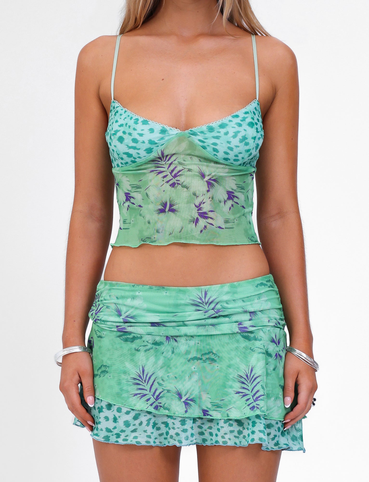 A person is wearing the TIGERMIST SAIGE TOP in green, which features a mixed pattern of leopard spots, palm leaves, and floral prints. The matching ensemble includes an adjustable elastic top that gives it a light, summery appearance. The individual has long blonde hair and is accessorizing with silver bracelets on both wrists.