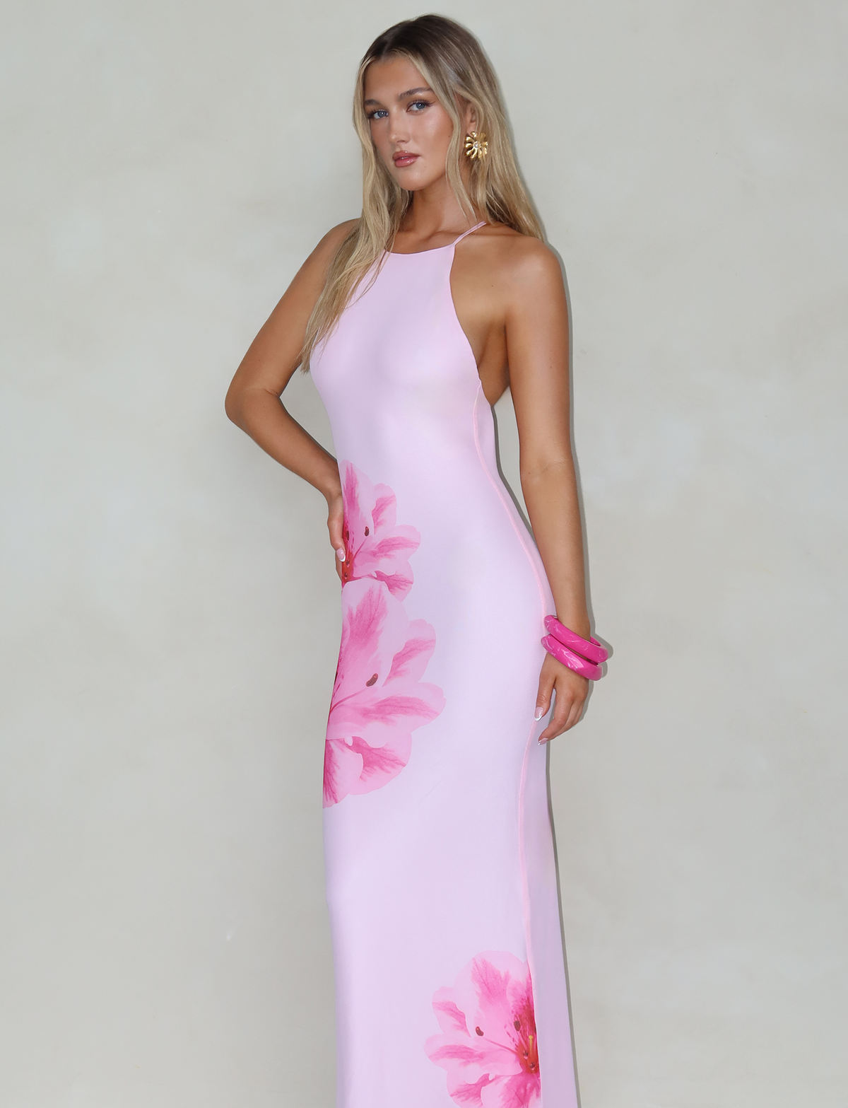 A woman stands against a plain background, dressed in the KARTER DRESS - PINK : BABY PINK by TIGERMIST, a sleeveless, form-fitting dress with mesh floral fabrication. She accessorizes with a pink bracelet and gold earrings, while her long blonde hair cascades down. She poses confidently with one hand on her hip.