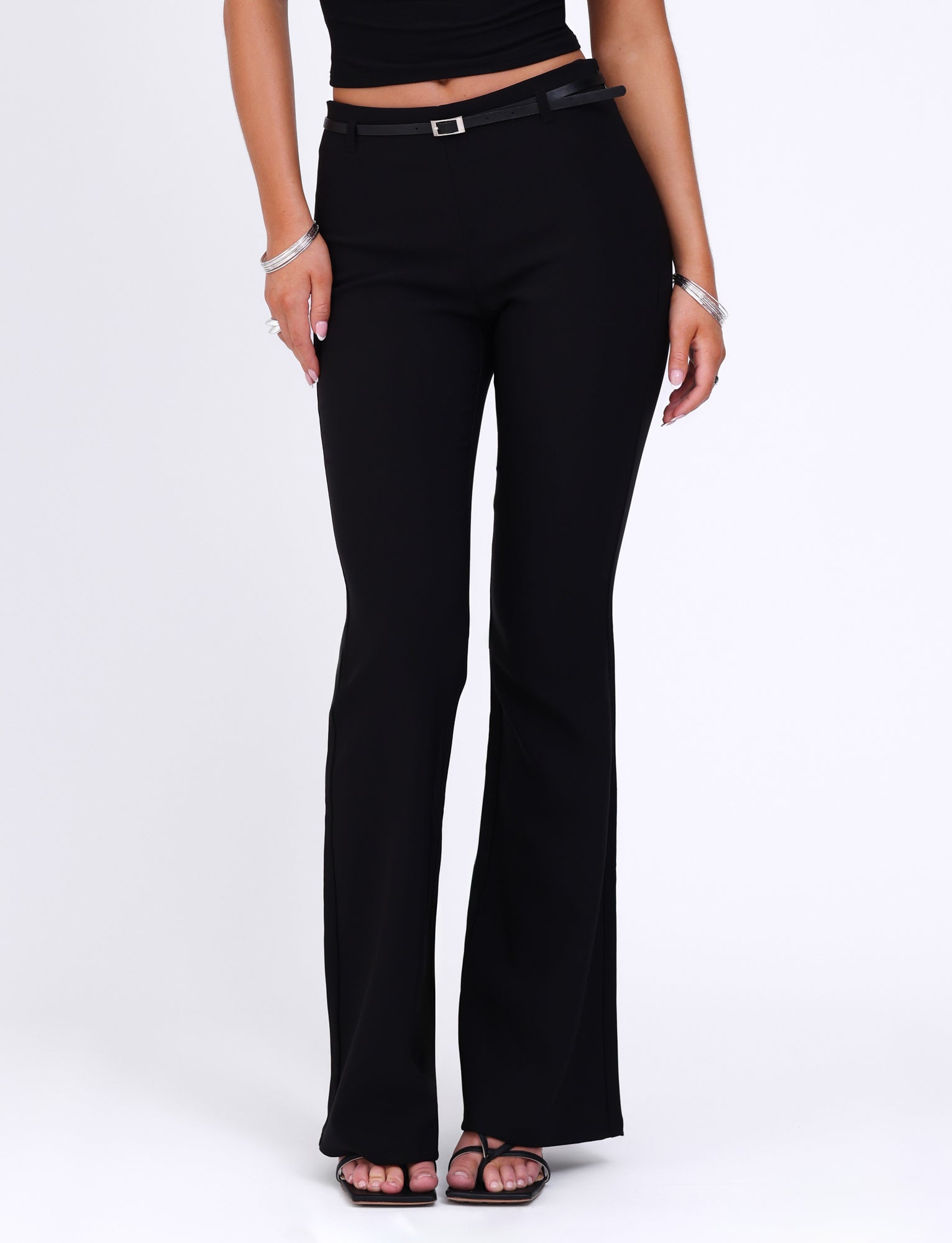 A person is wearing a black sleeveless top and the TIGERMIST TERLA PANT in black, which features a mid-rise waist and a flared silhouette. They stand with their hands relaxed at their sides, displaying silver bracelets on their left wrist. The flared pants complement their black strappy heels as they pose against a white background.