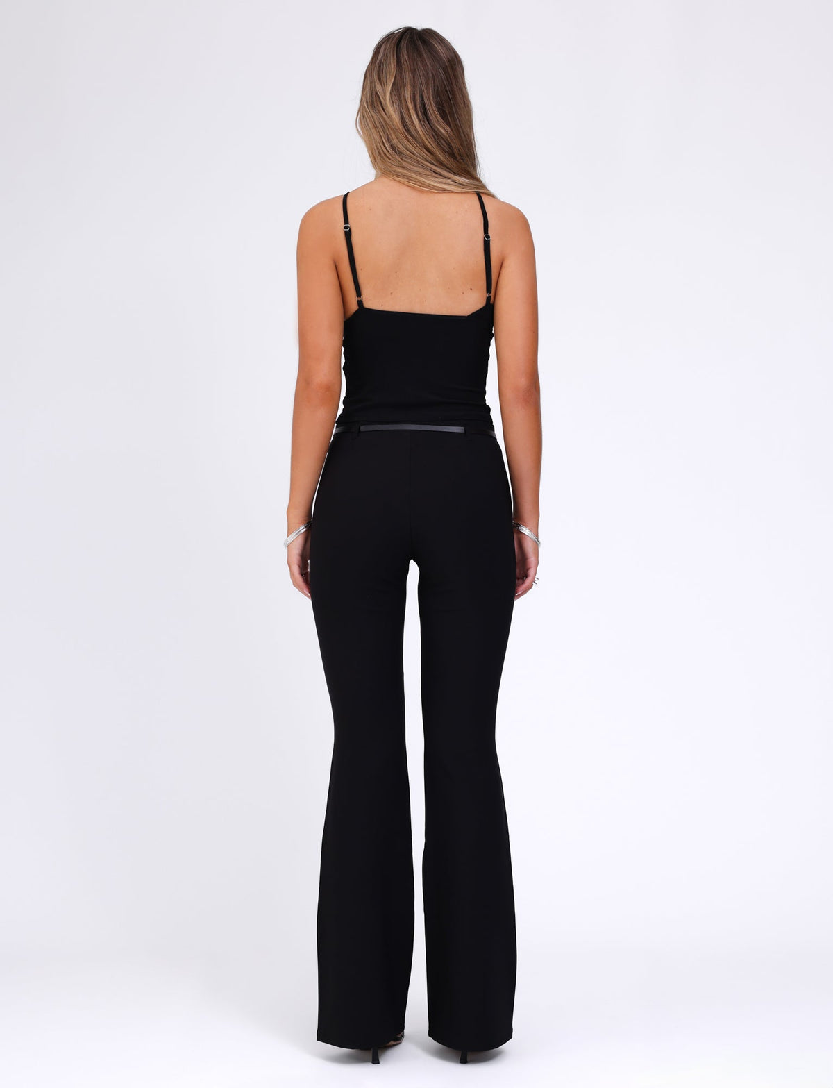 A person with long hair is standing with their back to the camera. They are wearing a black sleeveless top and the TERLA PANT - BLACK from TIGERMIST, featuring a mid-rise waist and a flared silhouette. The background is plain white.