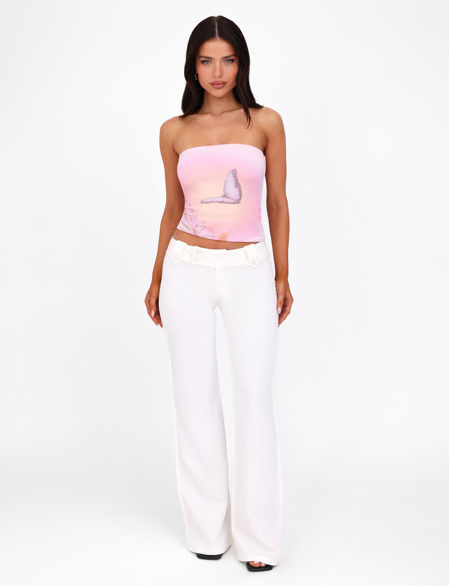 A woman with long dark hair stands confidently against a white background. She is wearing the TIGERMIST AMBROSE BOOB TUBE in pink, featuring an ombre butterfly graphic, paired with high-waisted white pants. Her hands are relaxed by her sides.