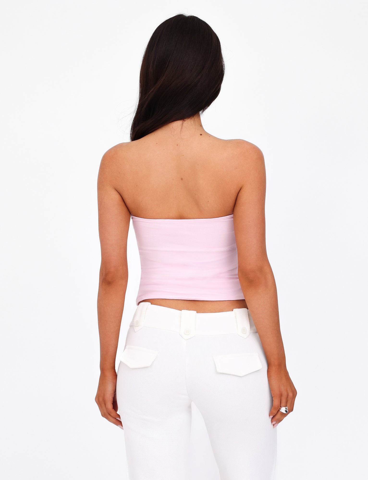 A woman with long dark hair is seen from behind, wearing the AMBROSE BOOB TUBE - PINK by TIGERMIST paired with high-waisted white pants featuring button details on the pockets. She stands against a plain white background that accentuates her style as effortlessly as a bodycon dress molds to form.