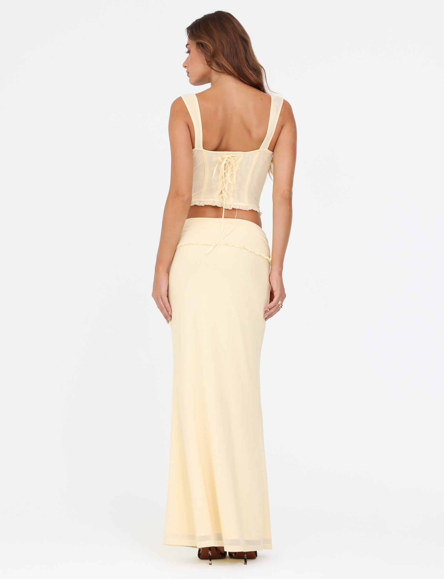 A woman stands with her back to the camera, wearing a cream-colored, sleeveless top featuring lace-up details paired with the TIGERMIST GINNY MAXI SKIRT in yellow lemon. Her long brown hair cascades down against the plain white background.