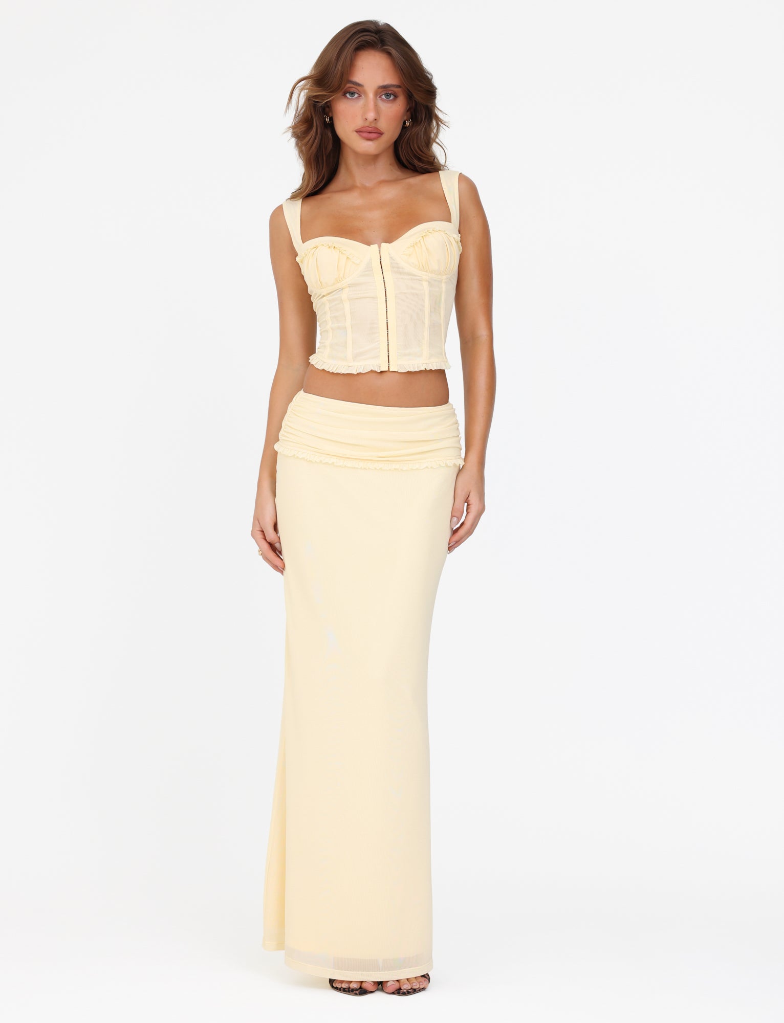 A woman poses confidently, standing against a plain white backdrop, wearing a light yellow corset top paired with the TIGERMIST GINNY MAXI SKIRT - YELLOW: LEMON, featuring its distinctive wide waistband. Her hair cascades loosely around her face as she radiates a serene confidence.