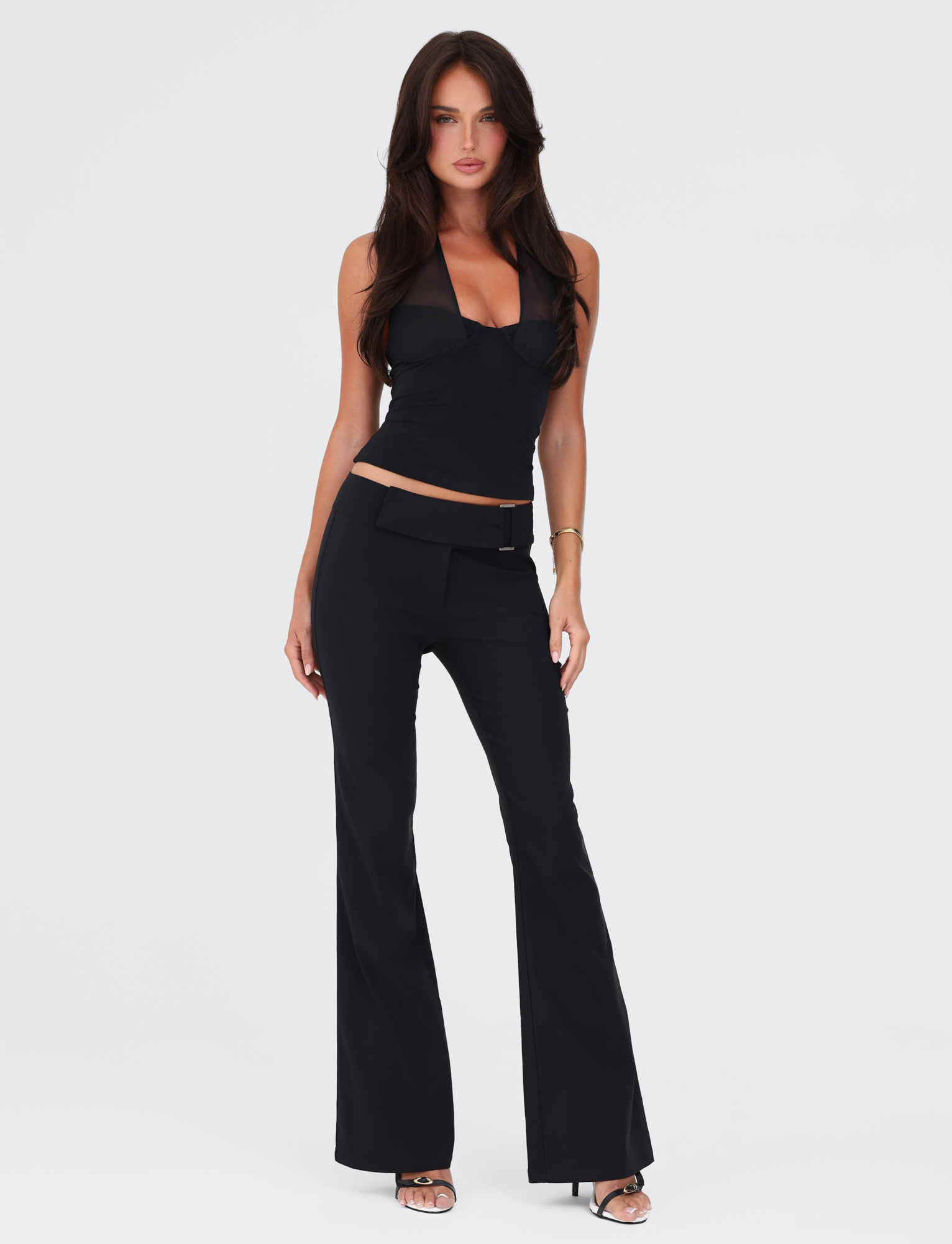A woman with long brown hair stands against a white background wearing the TIGERMIST GRANGER TOP in black, styled with black flared pants and silver open-toe heels. Her arms are relaxed at her sides and she has a neutral expression.