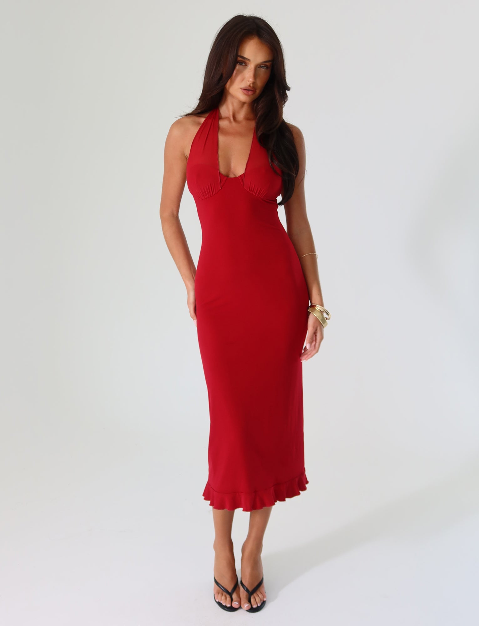 A woman wearing the TIGERMIST GRANGER MIDI DRESS in cherry red—featuring a fitted halter-neck design with ruffle details—poses against a plain background. The dress's flared hem is stylishly paired with black high-heeled sandals and gold bracelets on her wrist.