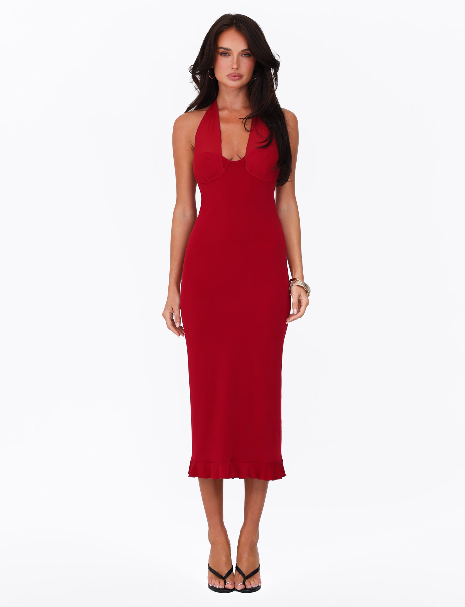 A person with long dark hair wears the sleeveless, fitted Granger Midi Dress in Cherry Red by Tigermist, featuring a ruffled hem, paired with black heels and standing against a plain white background.