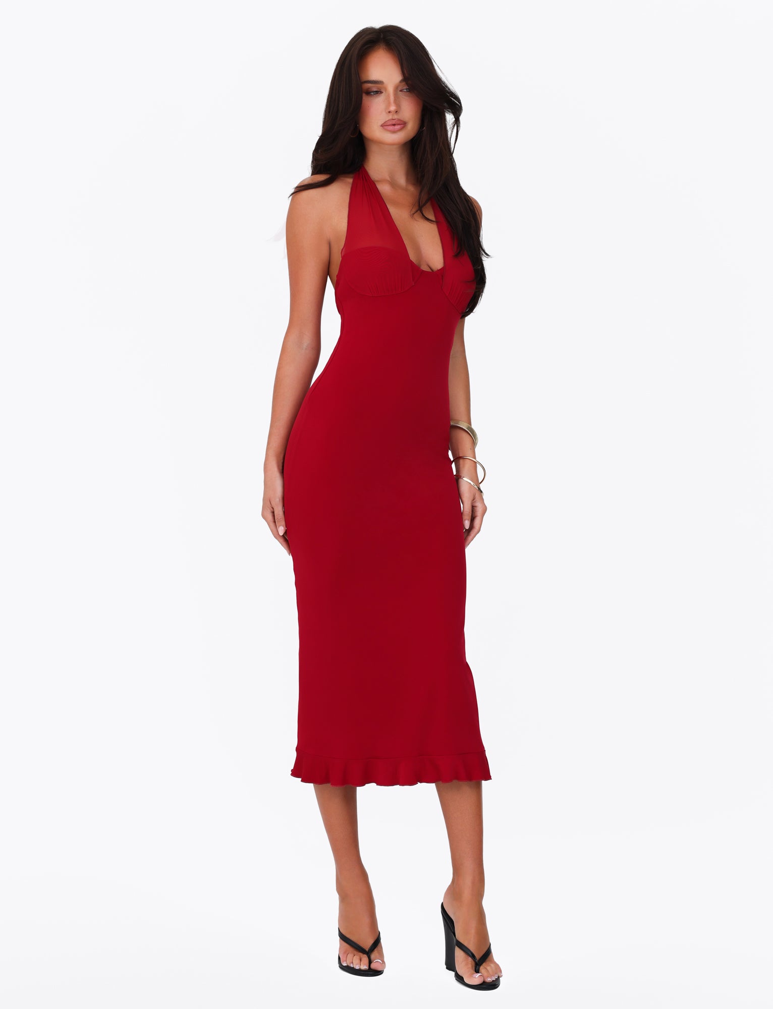 A woman poses against a plain white background wearing the Cherry Red GRANGER MIDI DRESS by TIGERMIST. The sleeveless halter dress features a ruffle detail and is fitted, extending to mid-calf with a slight frill at the hem. She completes her look with black high-heeled shoes, long dark hair, and bracelets on her left wrist.