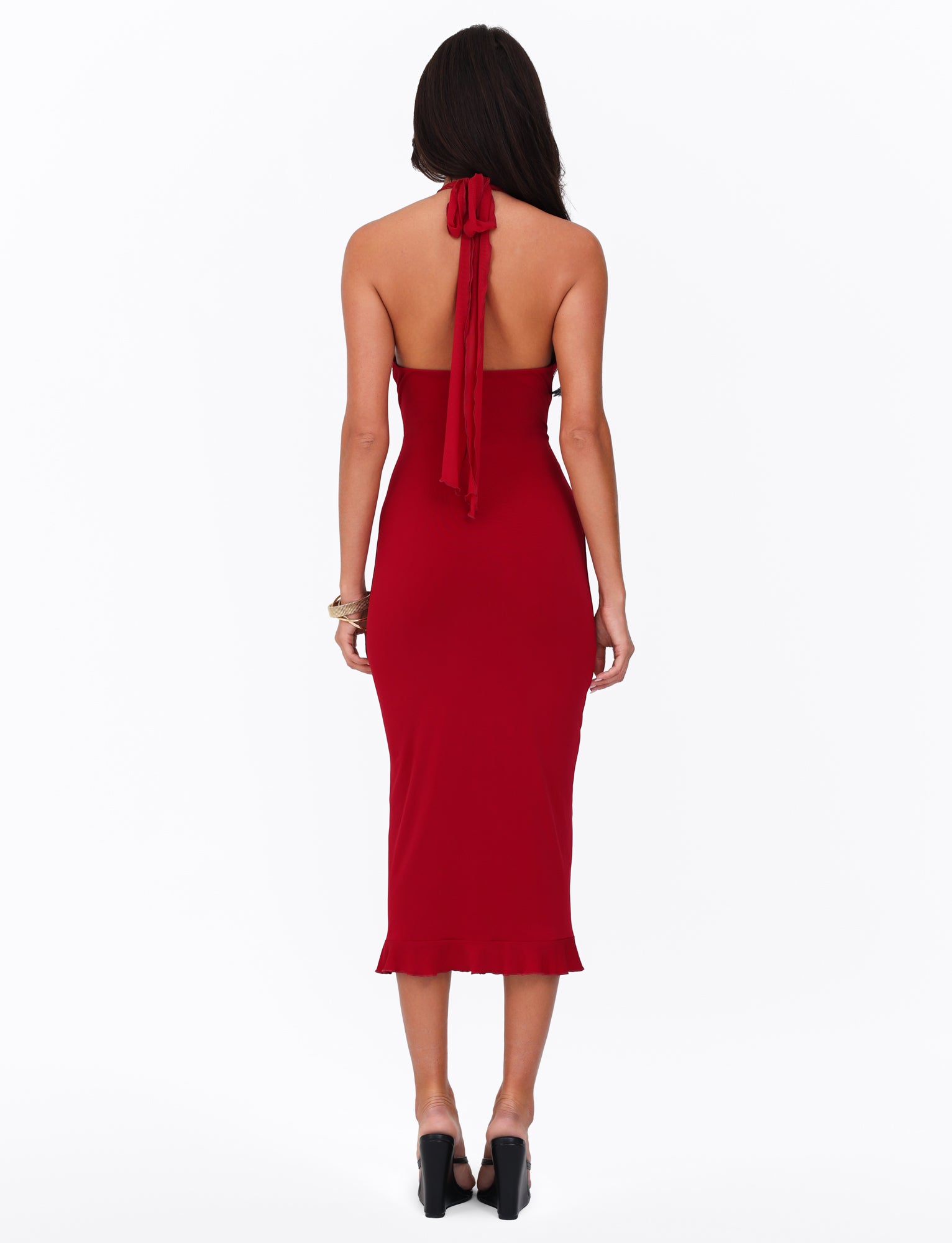 A woman with long dark hair stands with her back to the camera, wearing the GRANGER MIDI DRESS in Cherry Red by TIGERMIST. The sleeveless dress features a halter neckline and subtle ruffle detail, tying elegantly at the neck and falling gracefully to mid-calf. Her black high-heeled shoes complement the vibrant red dress against a plain white background.
