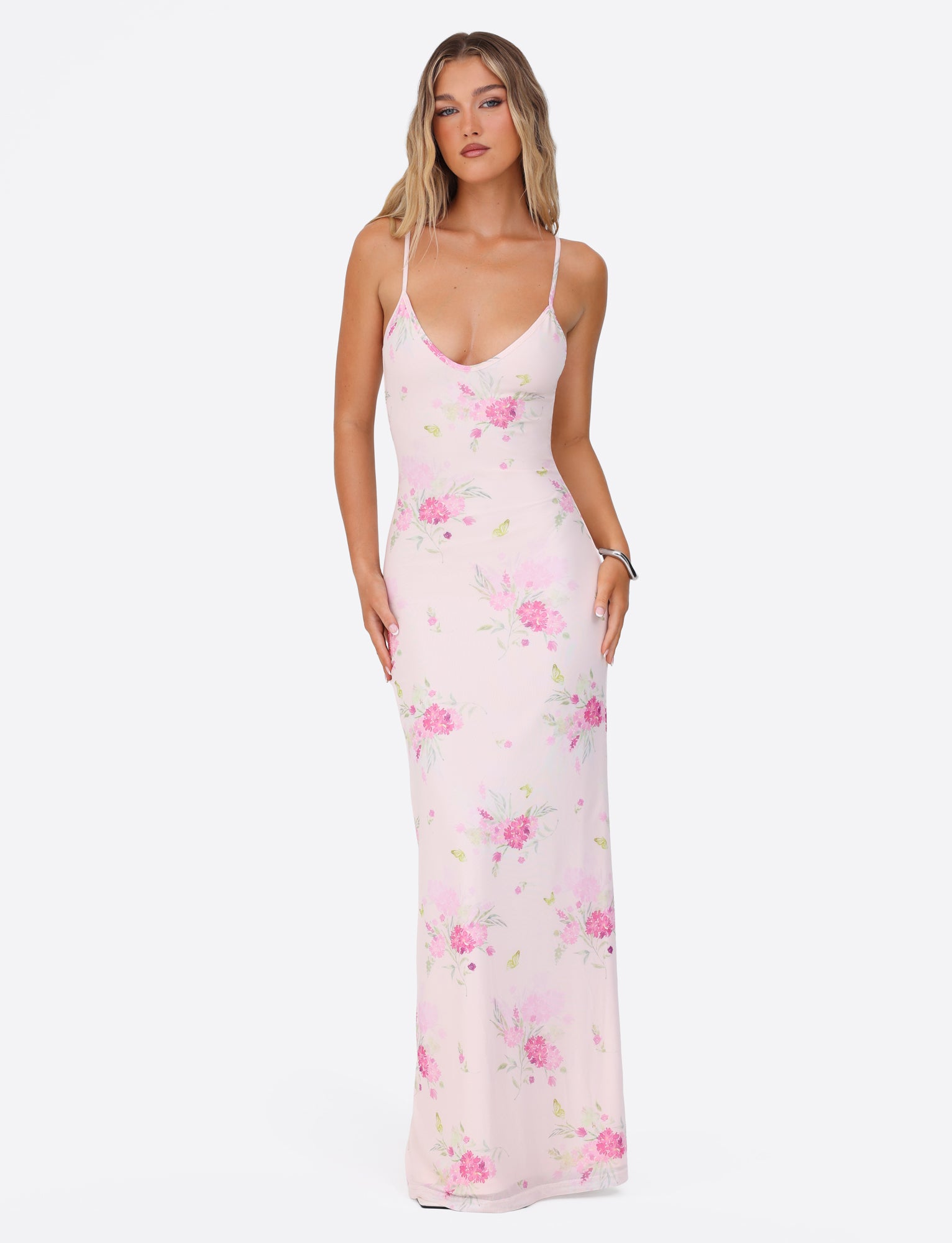 A woman wearing the OSIRIS DRESS from TIGERMIST, in a watercolor floral print of pink hues, stands against a white background. The dress, made with mesh fabric and adjustable spaghetti straps, has a long and flowing design. Her long hair cascades down as she gazes forward with a neutral expression.