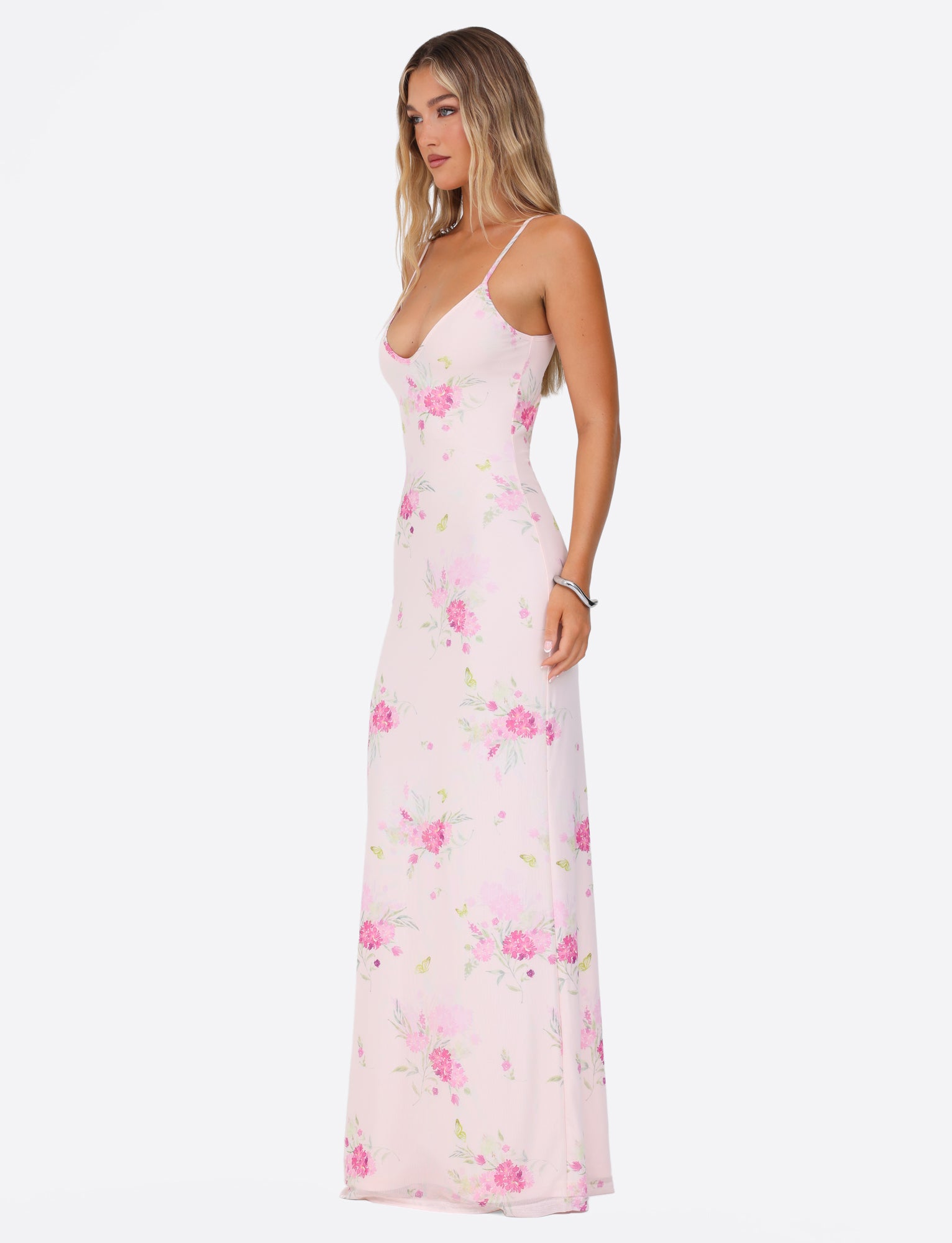 A woman models the OSIRIS DRESS by TIGERMIST, showcasing its light pink water colour floral design against a plain white background. The dress features adjustable spaghetti straps for a customized fit and an elegant, flowy silhouette. Her long hair cascades in loose waves.