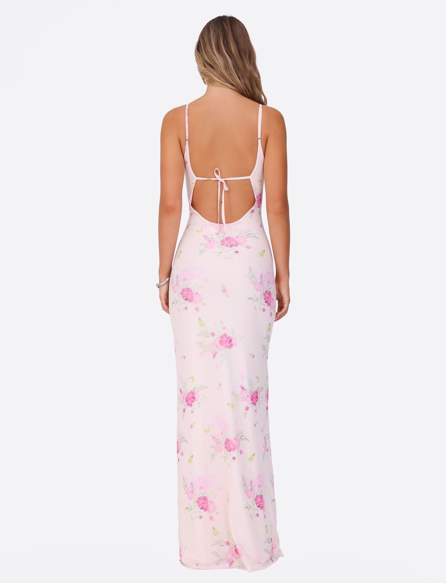 A woman with long hair is seen from the back wearing the TIGERMIST OSIRIS DRESS in a light pink, watercolor floral print. The dress, made of mesh fabrication, showcases a sleeveless design with an open back tied with a bow and features a flowing maxi length. She stands against a plain white background.