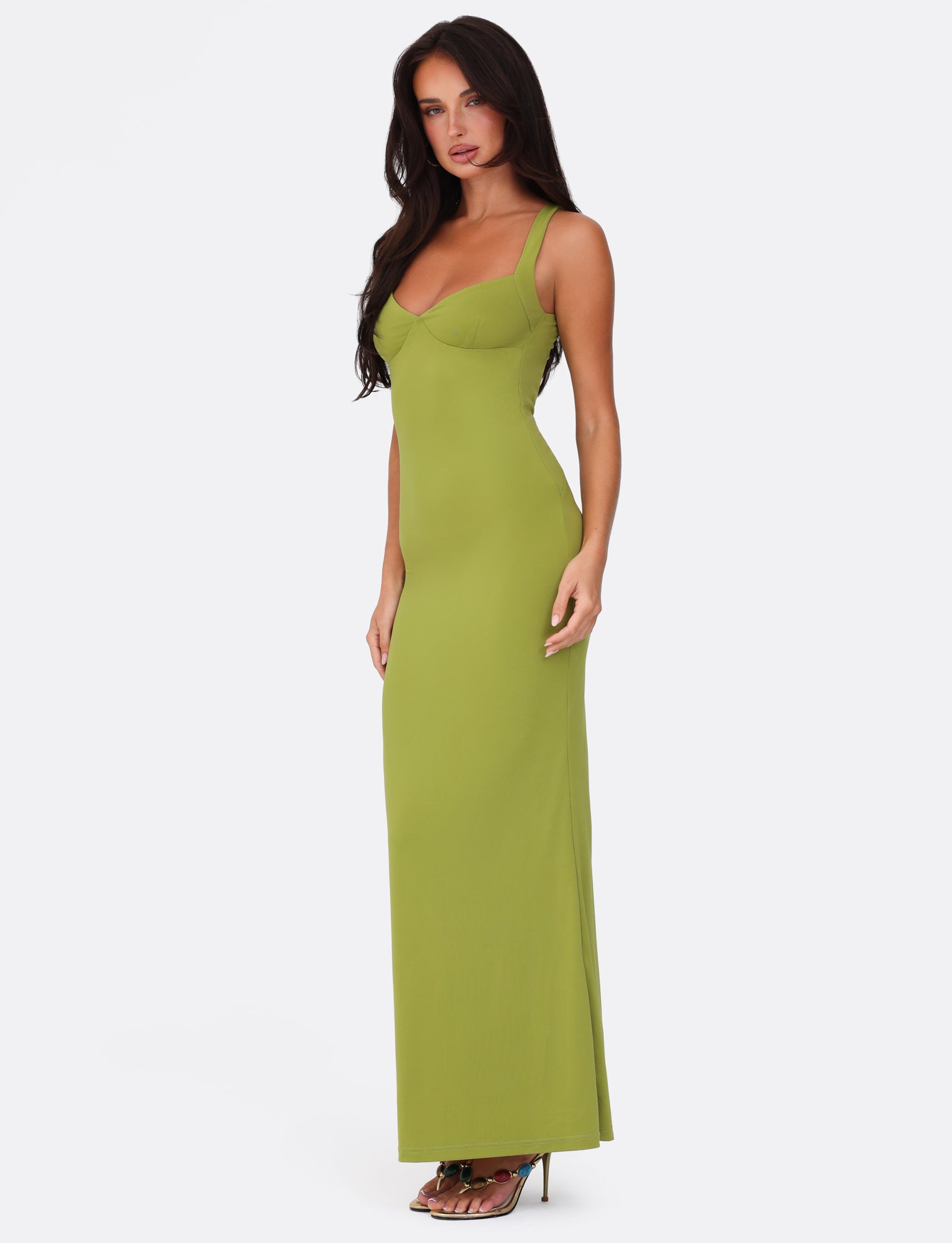 A woman in the SONNET MAXI DRESS by TIGERMIST, featuring a green color and a sweetheart neckline, stands against a plain white background. She has long dark hair and pairs the dress with high-heeled sandals, posing with her right hand resting on her hip.