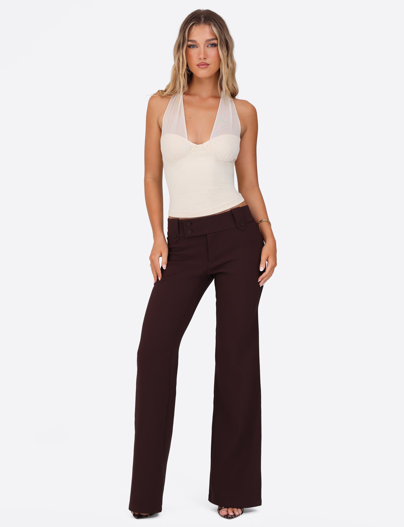 A person poses against a plain background, wearing a sleeveless Granger Top from Tigermist, featuring a halter neckline and pairing it with brown high-waisted, wide-leg pants. The outfit exudes style and elegance, creating a minimalistic fashion statement.