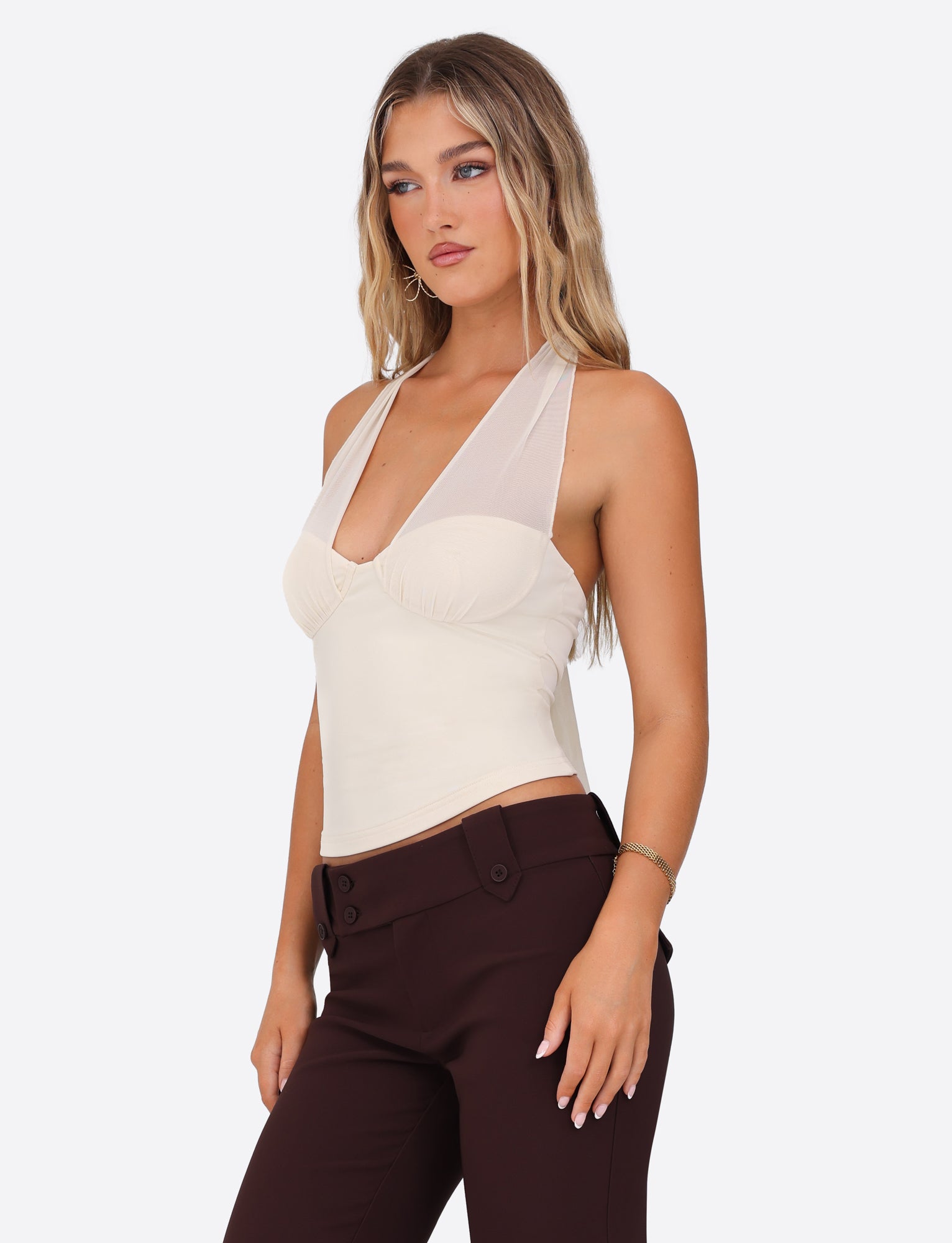 A woman with long blonde hair is wearing a sleeveless TIGERMIST Granger Top in cream, featuring a halter neckline, paired with dark brown pants. She accessorizes with hoop earrings and is gazing sideways against a plain white background.