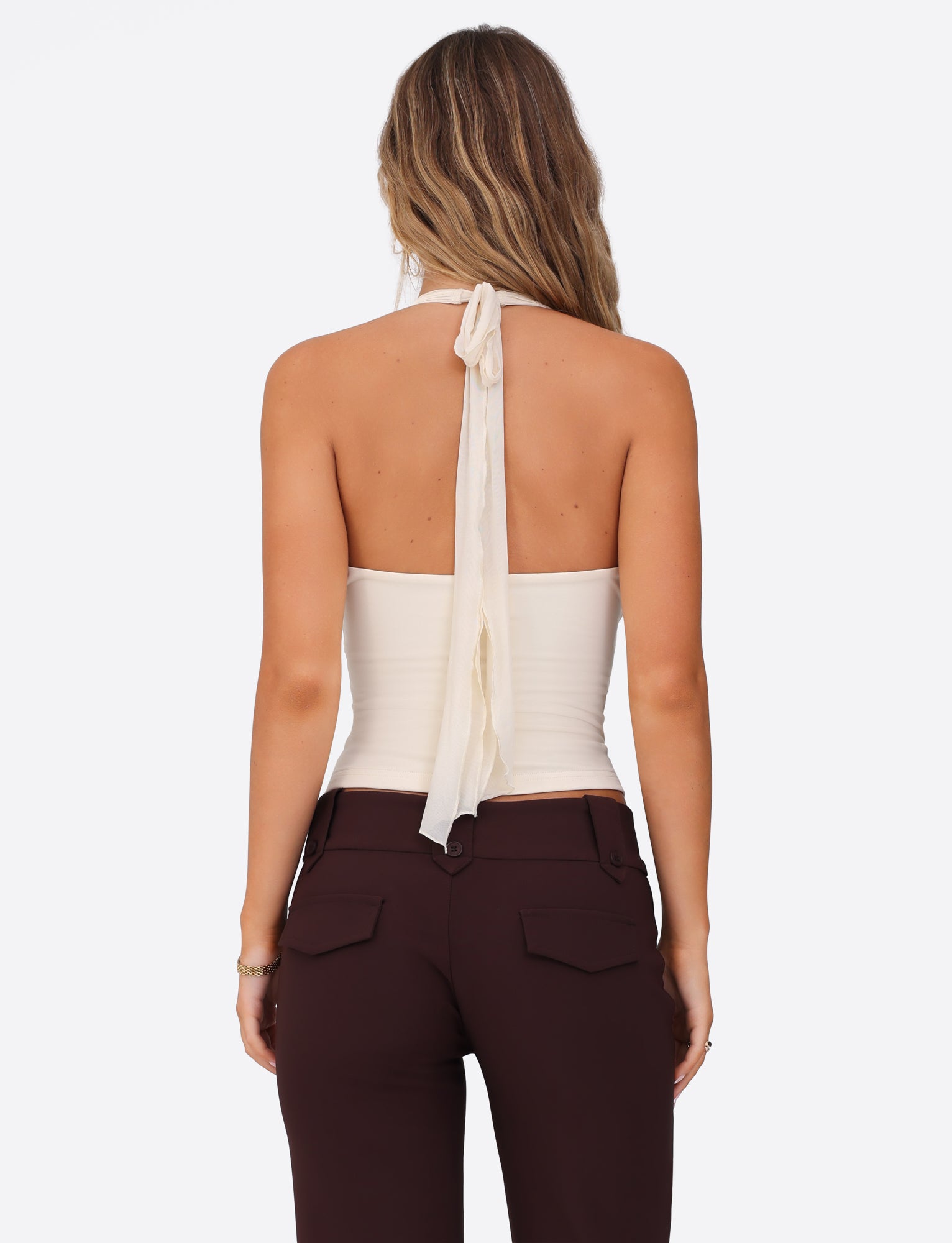 A rear view captures a person with long wavy hair dressed in a TIGERMIST Granger Top, available in white and cream, featuring a halter neckline, paired with brown high-waisted pants against a plain white background.