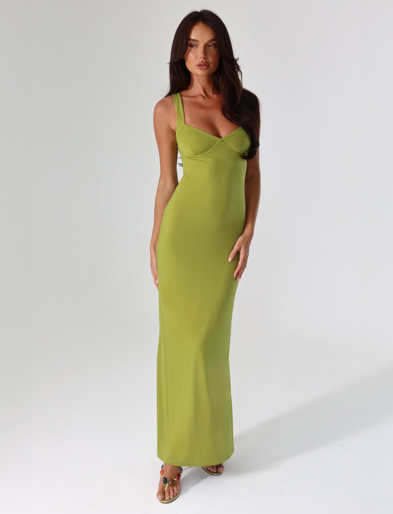 Dressed in the SONNET MAXI DRESS by TIGERMIST, a person strikes a pose on a plain white background. The green dress features a form-fitting, maxi-length design with a sweetheart neckline. Complementing their long dark hair are open-toed sandals, and their hands rest gently on their hips, exuding an air of elegance and style.