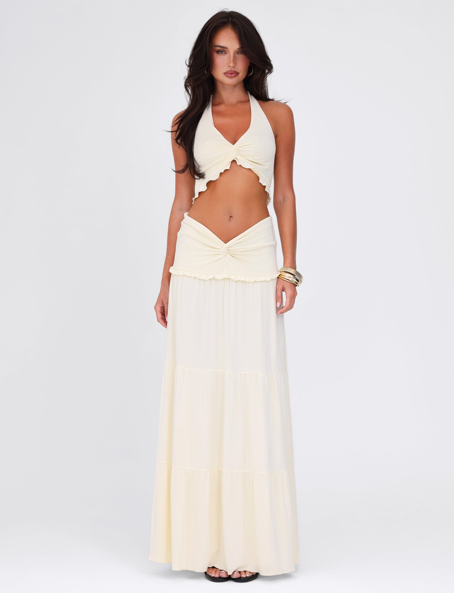 A confident woman poses against a plain backdrop wearing the KATYA HALTER TOP - WHITE : CREAM by TIGERMIST, featuring a twist detail and cropped length, paired with a matching long skirt and accessorized with bracelets.
