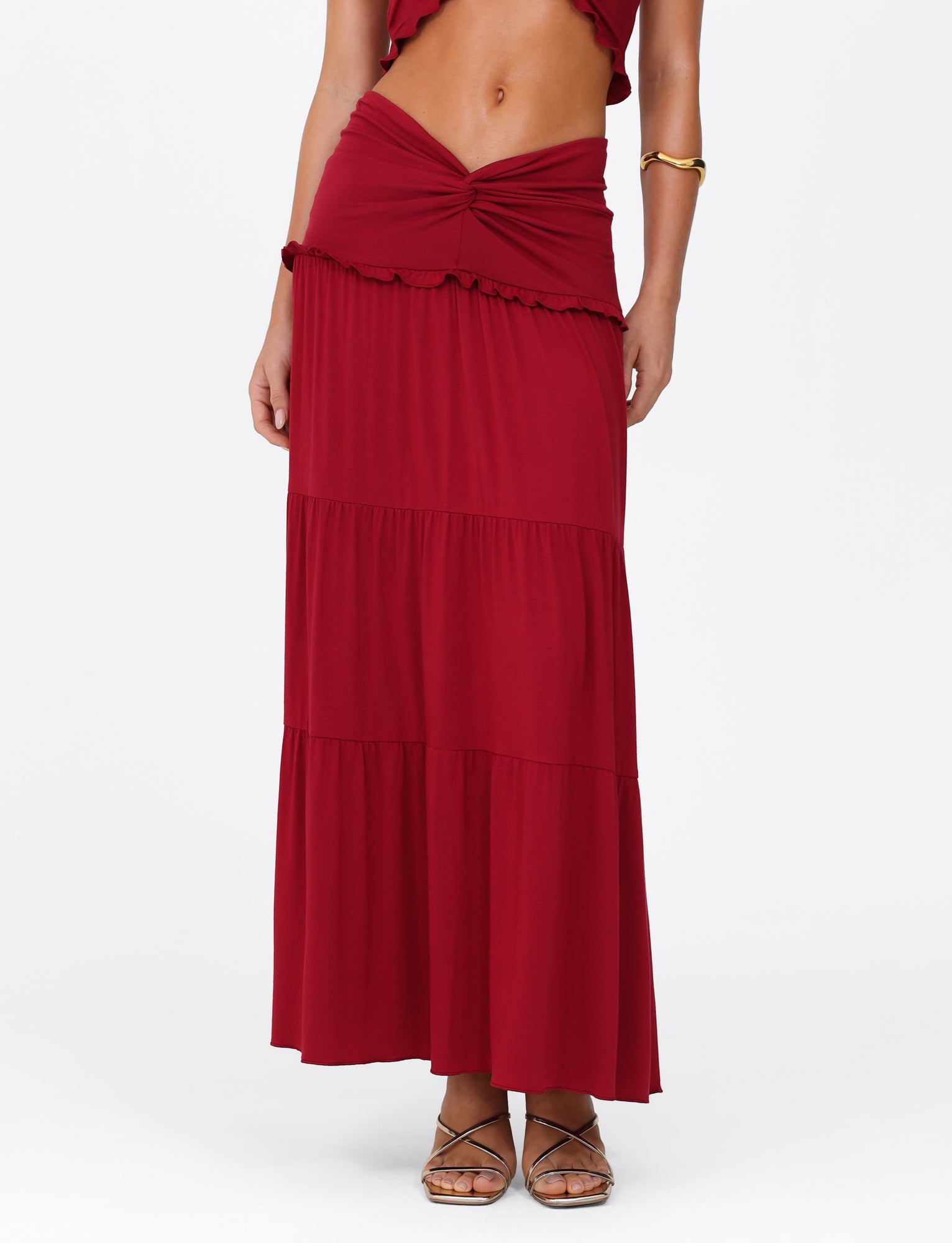 A person is dressed in the TIGERMIST KATYA MAXI SKIRT - RED, featuring tiers and a wide waistband with a knot detail, paired with strappy sandals and a gold bracelet on the left wrist. The image is cropped above the shoulders against a white backdrop.
