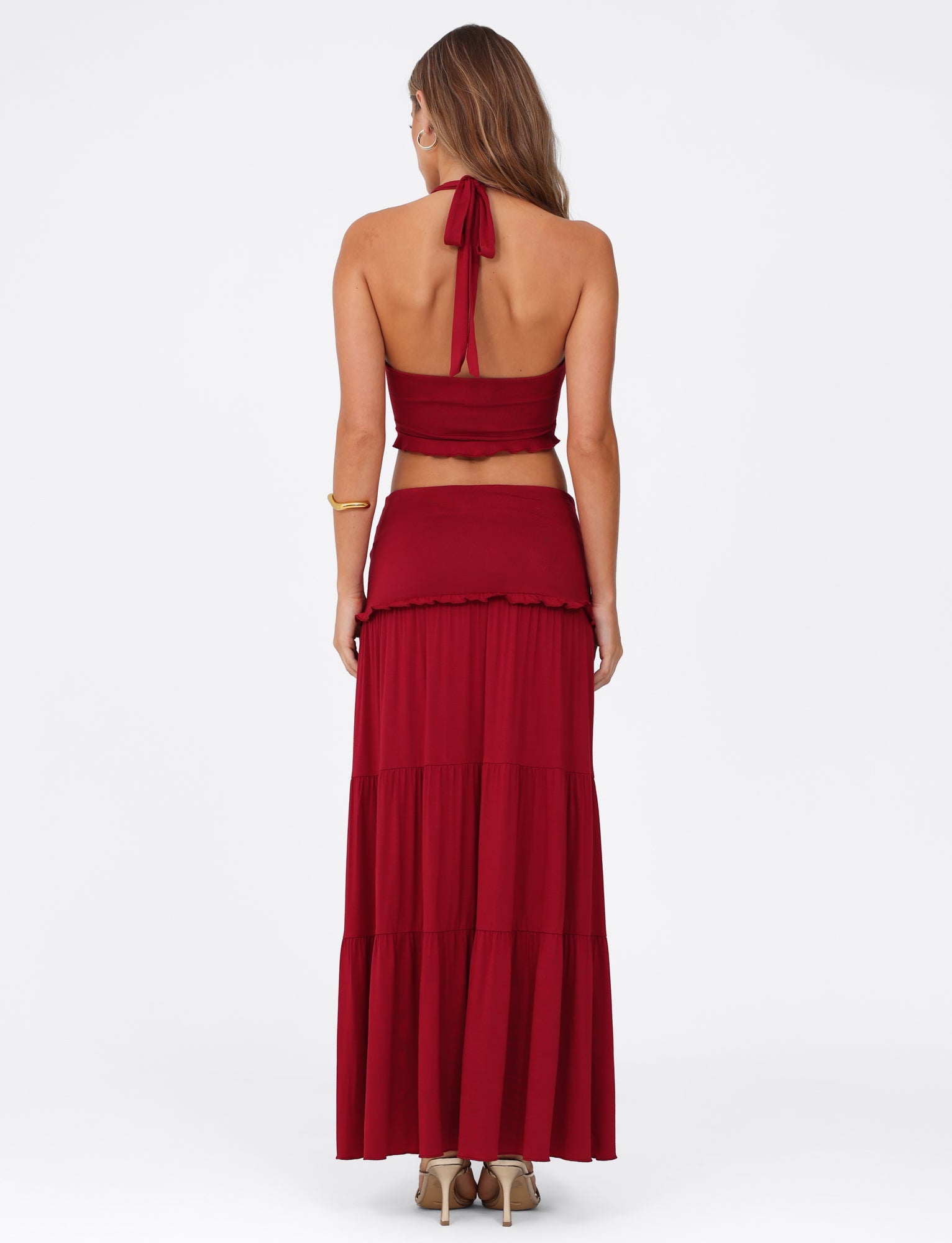 A woman showcases the TIGERMIST KATYA MAXI SKIRT - RED with its wide waistband and tiered design. Her flowing hair pairs beautifully with high-heeled sandals, all captured against a plain white background.