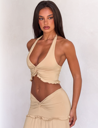 A woman with long dark hair models the TIGERMIST KATYA TOP in tan, styled as part of a matching set with a high-waisted skirt. She stands against a simple light background, gazing confidently at the camera.