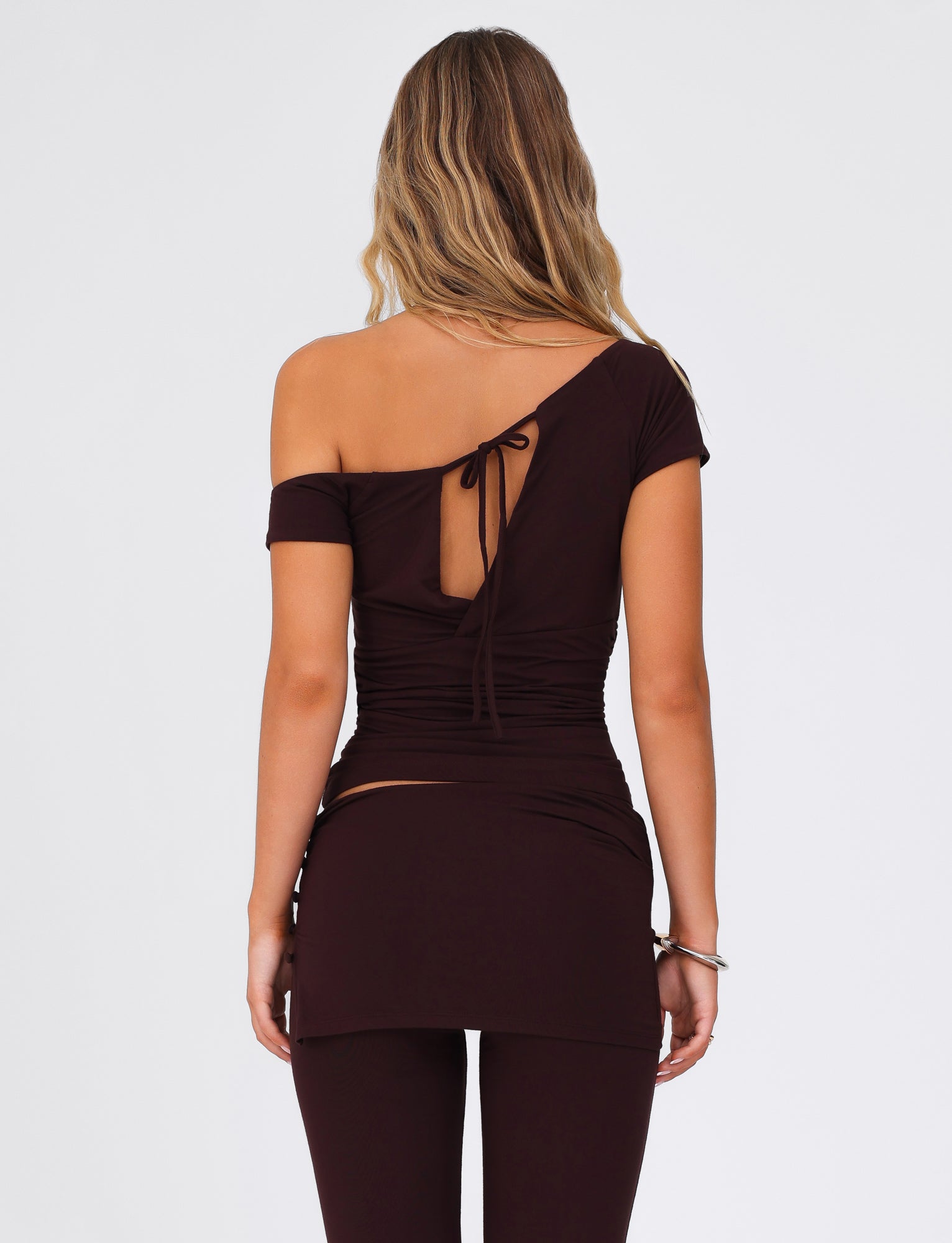A person with long hair is seen from the back wearing the LEILANI TOP - BROWN : CHOCOLATE by TIGERMIST. This buttery jersey top features an off-shoulder design and a back tie, paired with matching leggings, set against a plain white background.