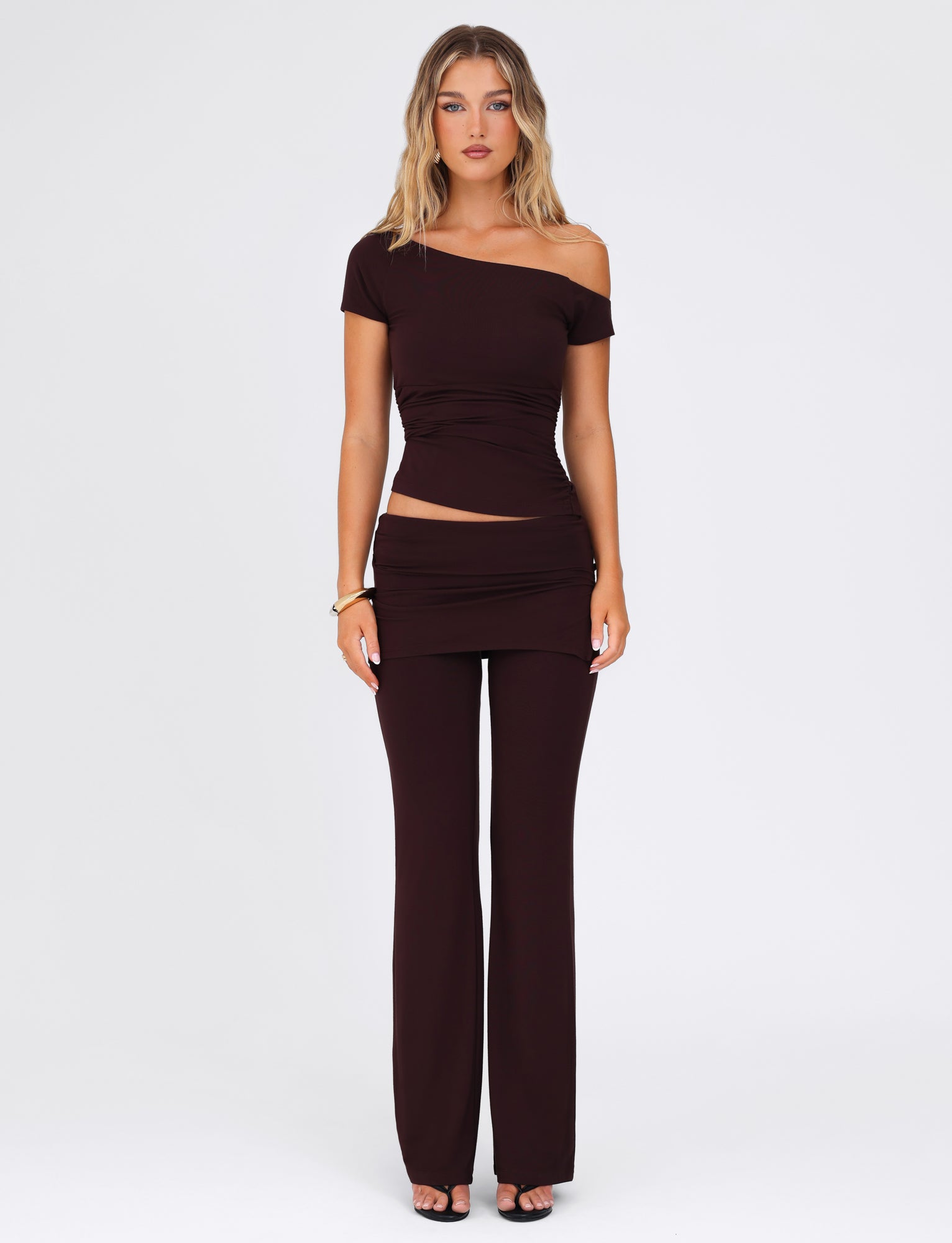 A person wearing a TIGERMIST off-shoulder dark brown top and the matching LEILANI PANT - BROWN: CHOCOLATE stands against a plain white background. They have light brown hair, wear black heels, and accessorize with a bracelet on one wrist.