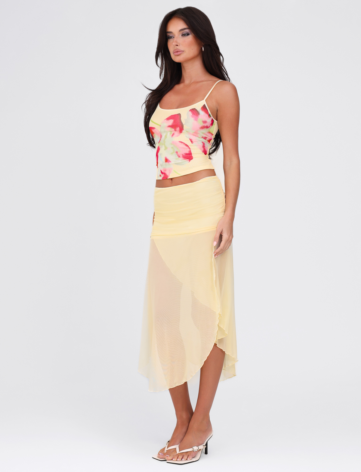 A woman with long dark hair wears a colorful tank, the TIGERMIST MARIANA SKIRT in pale yellow mesh, and white heeled sandals while posing against a plain white background.