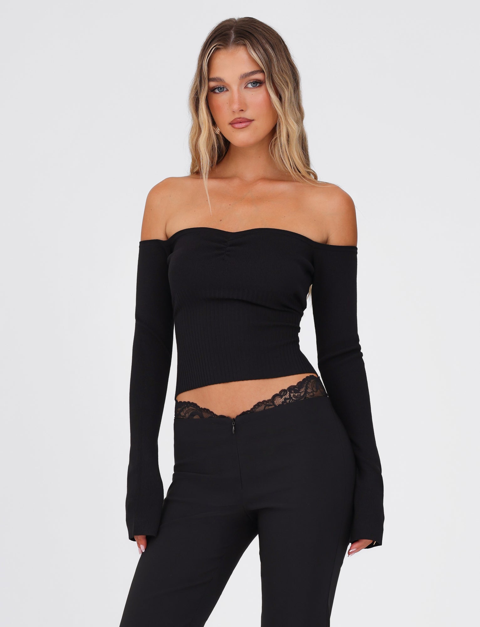 A woman with long, wavy hair wears the chic LILYANA LONG SLEEVE TOP - BLACK by TIGERMIST, featuring rib detail and paired with black pants. She stands against a plain white background, looking directly at the camera with her hands confidently on her hips.