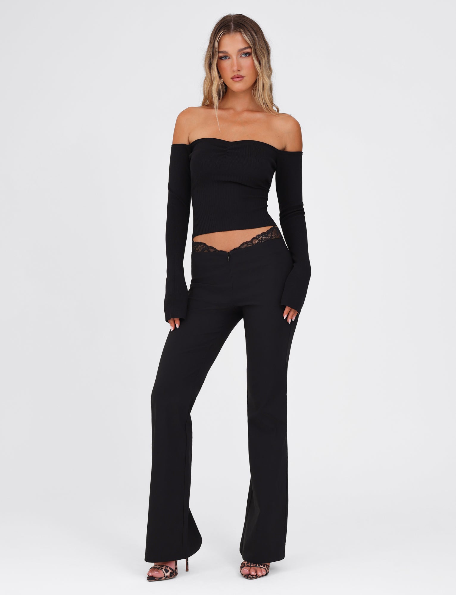 A woman dressed in the NYOMI PANT in black by TIGERMIST stands against a plain white background, wearing a fitted black off-shoulder top with flared leg pants adorned with black lace detail and open-toe heels.