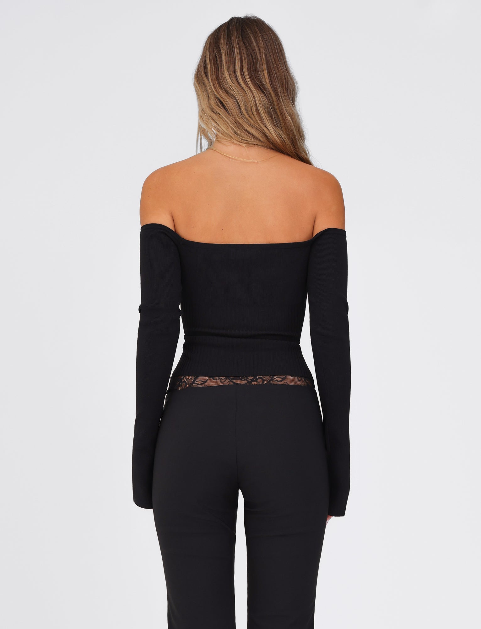 A woman with long hair faces away, dressed in the TIGERMIST LILYANNA Long Sleeve Top in black and black pants featuring a lace waistband, set against a plain white background.