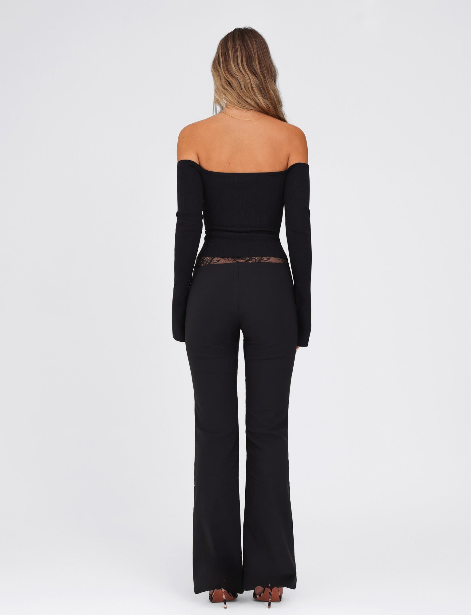 In front of a plain white background, a person with long hair stands facing away, wearing the off-the-shoulder black Nyomi Pant by TIGERMIST, featuring a matching top and flared leg design in high stretch fabric.