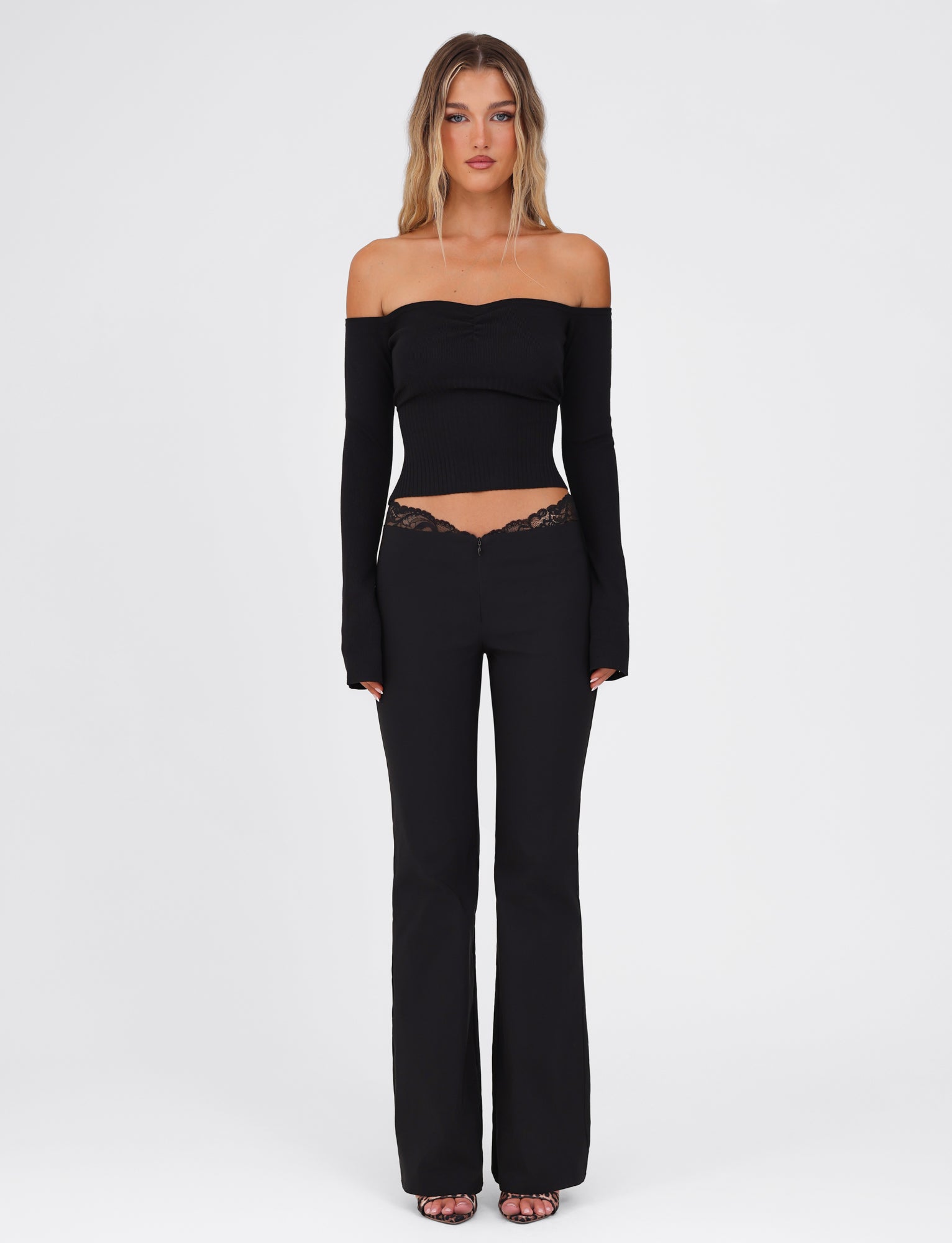 A woman stands against a plain white background, wearing a black, off-the-shoulder top with long sleeves paired with the NYOMI PANT - BLACK from TIGERMIST, featuring high stretch fabric and lace details. Her long, wavy hair cascades down as she completes the look with stylish sandals.