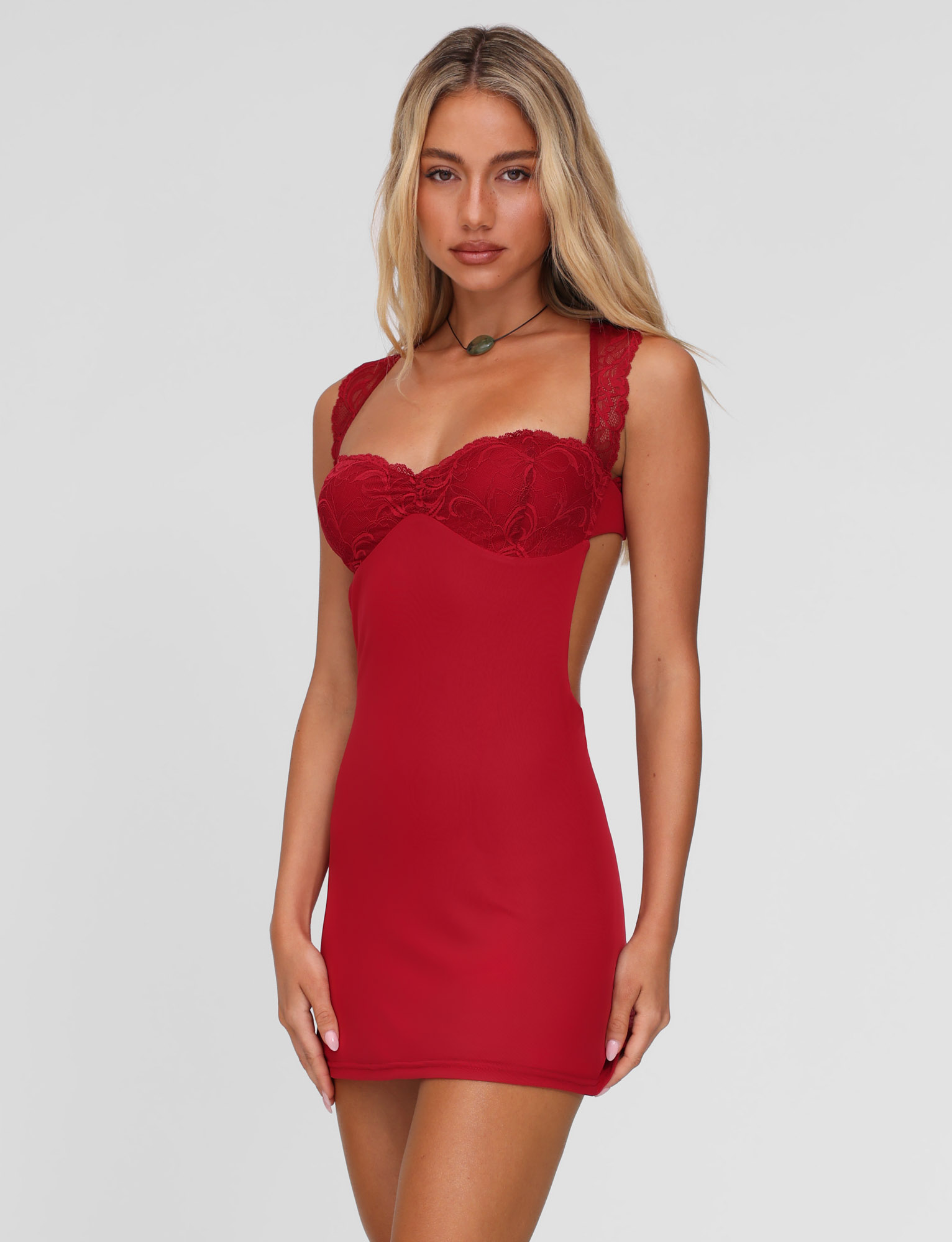 A woman with long blonde hair wears the TIGERMIST NYOMI MINI DRESS in red, styled with a green pendant necklace, standing against a plain light grey background.