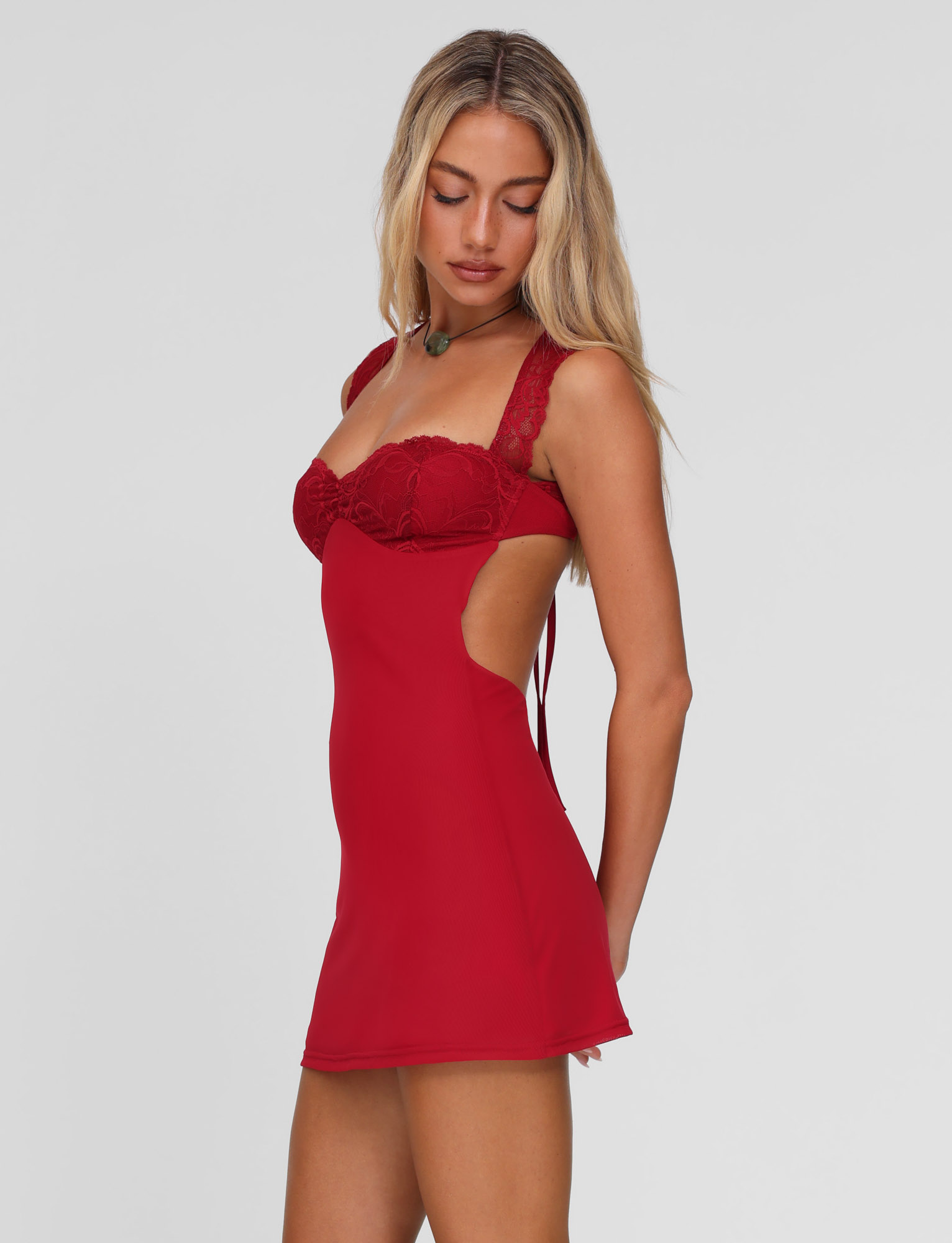 A woman with long blonde hair models the TIGERMIST NYOMI MINI DRESS - RED, showcasing its lace bust panel and open back, against a plain light gray background.