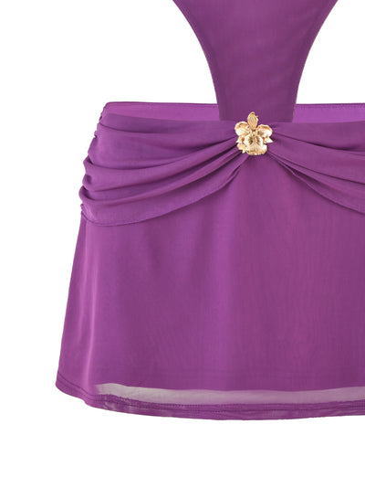 A close-up of the TIGERMIST KYNLEE MINI DRESS - PURPLE showcases its fitted bodice with draped fabric around the waist. The dress features a gold and white floral brooch at the center of the waistline. The skirt portion has a slightly flared design with a sheer overlay at the bottom, adding an elegant touch.