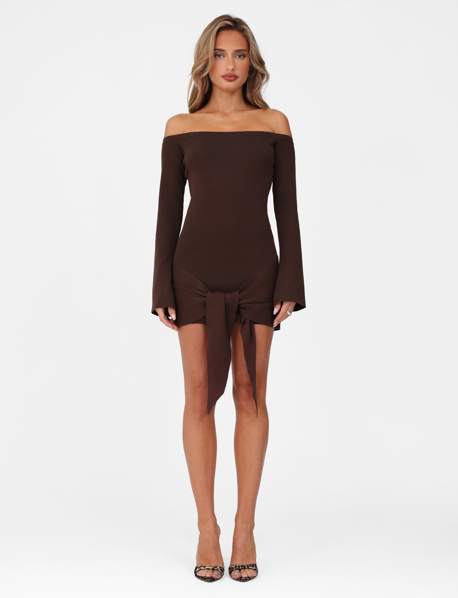 A woman poses against a white background in the TIGERMIST CYPRESS MINI DRESS - BROWN : CHOCOLATE, an off-the-shoulder, bodycon dress with a front tie detail. She pairs it with black open-toe heels and wears her hair loose and wavy.
