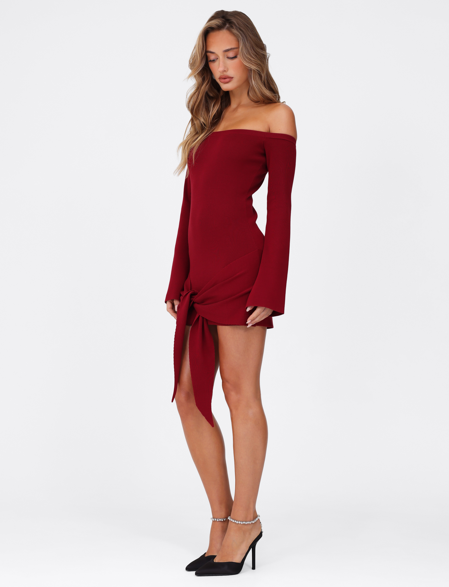 A woman stands sideways in the TIGERMIST CYPRESS MINI DRESS - RED : WINE, a bodycon off-the-shoulder mini with long sleeves and front tie detail, paired with black pointed ankle-strap heels against a plain white background.